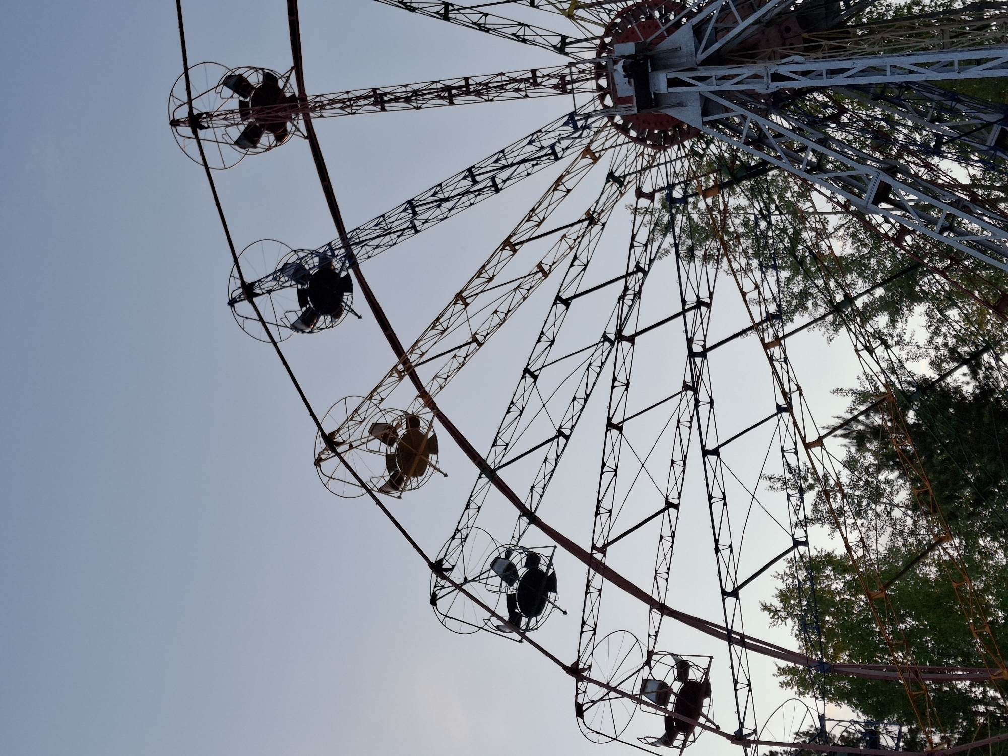 The image shows a Ferris wheel in Chaikovsky, Russia, viewed from a low angle, pointing upwards. The wheel’s structure is made of metal, with a combination of red, white, and black elements. The individual cars of the Ferris wheel are simple metal cages. Some cars appear to be occupied by people. The sky is visible in the background, along with some foliage of trees. The image's perspective emphasizes the height and structure of the Ferris wheel.