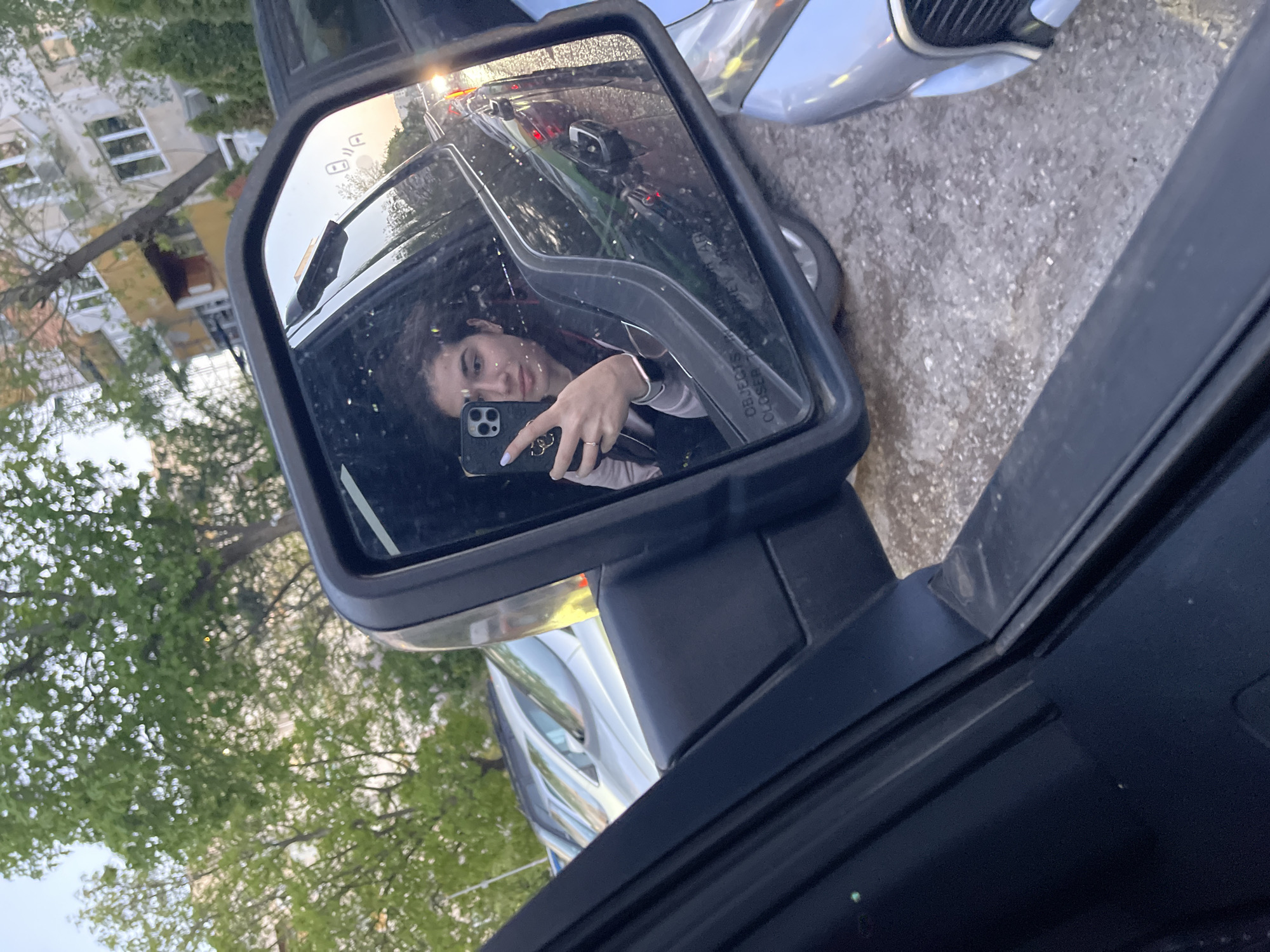 A photograph taken from inside a vehicle shows the driver's side mirror reflecting a young woman holding a smartphone in her right hand, presumably taking a selfie. The reflection in the mirror also includes a portion of the adjacent car, the street, and a building across the street. The location appears to be an urban street in Sofia, Bulgaria, based on the style of the architecture and vehicles. The photograph is taken at dusk.