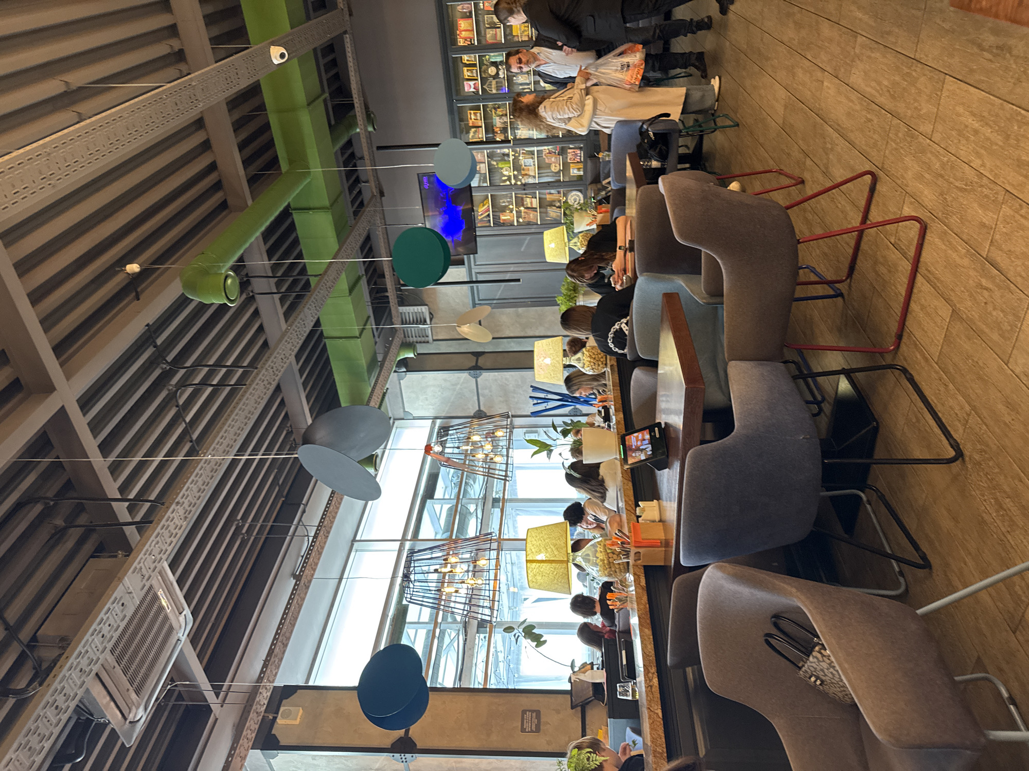 The image captures the interior of a restaurant in Saint Petersburg, Russia. Several patrons sit at a long communal table, some engaged with mobile devices. Large windows provide natural light, and pendant lights hang overhead. The restaurant features an industrial aesthetic with exposed ceiling beams, ductwork painted green, and air conditioning units. The chairs are a mix of gray and brown upholstery with metal frames, some with red accents. In the background, a display cabinet is filled with various items. Other customers are visible near the display.