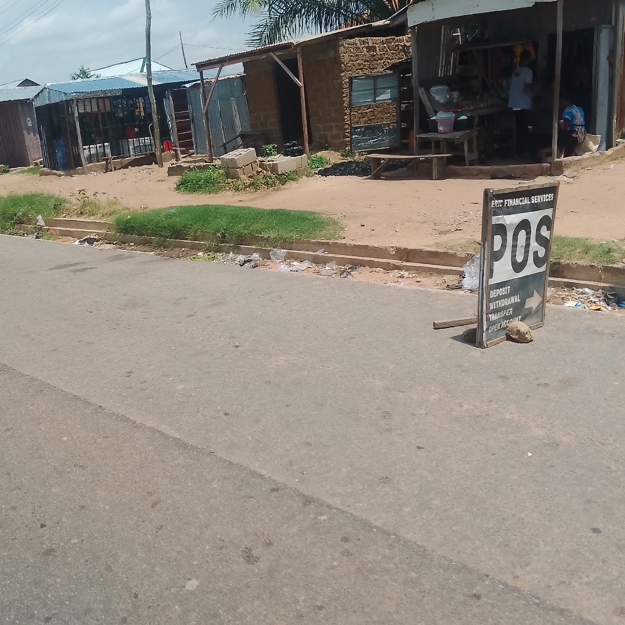 The image depicts a street scene in Jalingo, Nigeria. The foreground is dominated by an asphalt road, showing some wear and tear. To the right of the road, a sign for "ERIC FINANCIAL SERVICES" advertising "POS" services, along with options for "DEPOSIT," "WITHDRAWAL," and "TRANSFER," is prominently displayed. The sign is propped up by a piece of wood and a rock. Beyond the sign, there are several small shops constructed with corrugated metal and brick. These shops appear to be part of a market or commercial area. In one of the shops, two individuals are visible; one standing and one seated. The background shows some vegetation, including grass and palm trees, and a cloudy sky. The general environment appears to be that of a developing urban or semi-urban area with modest infrastructure. There is some scattered litter along the roadside, indicating potential waste management challenges.