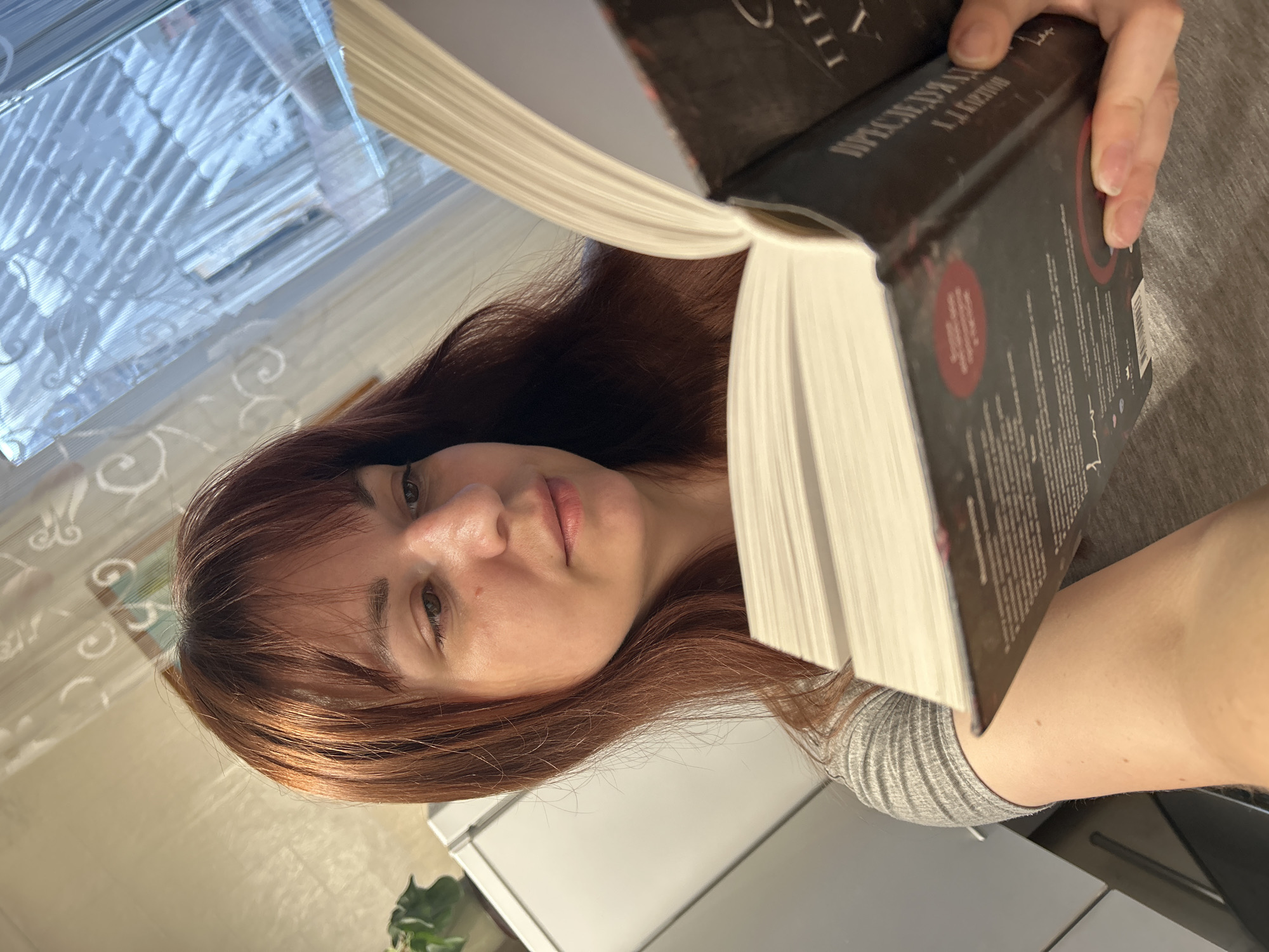 A woman with auburn hair holds open a thick book close to her face. The book cover is dark and partially obscures her face. The spine of the book has text in a Cyrillic alphabet. She wears a gray shirt. The background shows part of a window with patterned curtains, a white cabinet, and a potted plant. The location is not discernible as the city of Фоки, Russia based solely on the image.