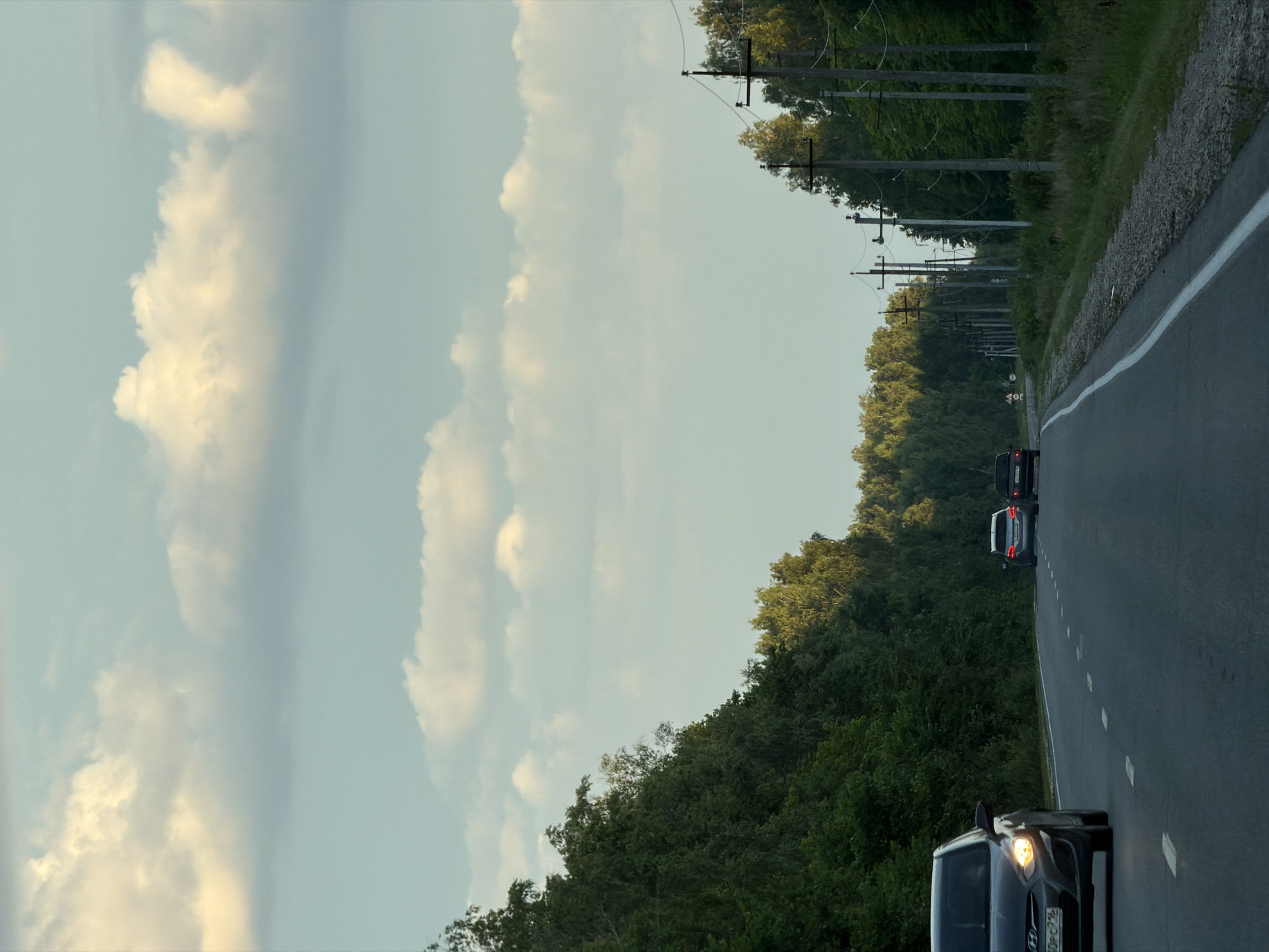 An image captures a paved road in Laishevo, Russia, bordered by dense green trees on the left and power line poles and vegetation on the right. The sky above is a clear, pale blue with scattered, fluffy white clouds, some of which are illuminated by warm, golden light, suggesting either early morning or late afternoon. Three cars are visible on the road: a dark grey Hyundai Solaris in the foreground on the left, its headlights on; a light-colored sedan further down the road; and a dark sedan behind it. The road has dashed white lane markings. The overall scene depicts a rural or semi-rural setting in Laishevo, Russia, under a partly cloudy sky.