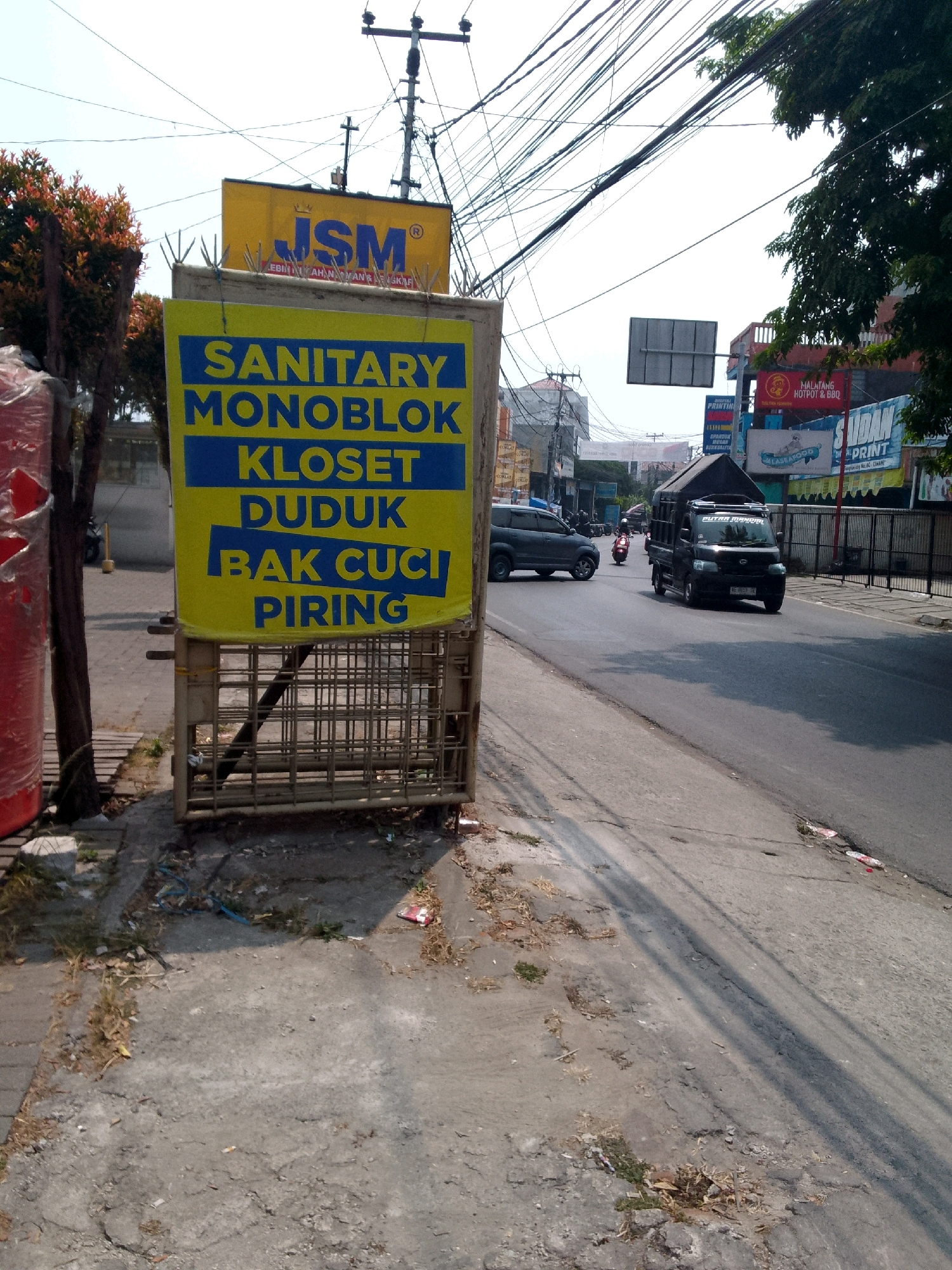 A yellow sign with blue text reads "SANITARY MONOBLOK KLOSET DUDUK BAK CUCI PIRING" and is mounted on a bamboo-like structure on the side of a road in Cimahi, Indonesia. The sign appears to be advertising bathroom fixtures. In the background, several vehicles, including a black truck and a dark SUV, are visible on the road. Utility poles and a tangle of overhead wires dominate the sky. Buildings and other signs are visible along the street. The foreground shows a cracked concrete sidewalk with some dry vegetation and debris. The overall scene depicts a commercial area on a sunny day.