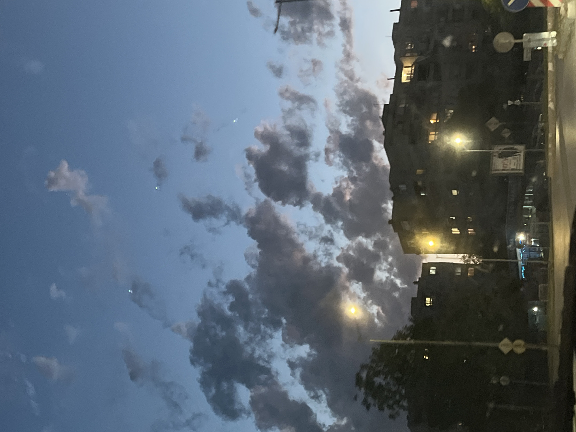 The image depicts a street scene in Shumen, Bulgaria, at dusk. In the foreground, there is a road with streetlights illuminating the area. The sky is a deep blue with scattered clouds, some of which are dark and ominous. Two streetlights are visible, one on the left side of the image and another further to the right. In the background, several multi-story apartment buildings are visible. The windows of these buildings are lit, indicating that people are inside. Some of the buildings appear to be older, with a Brutalist architectural style. The overall impression is one of a quiet, urban evening.