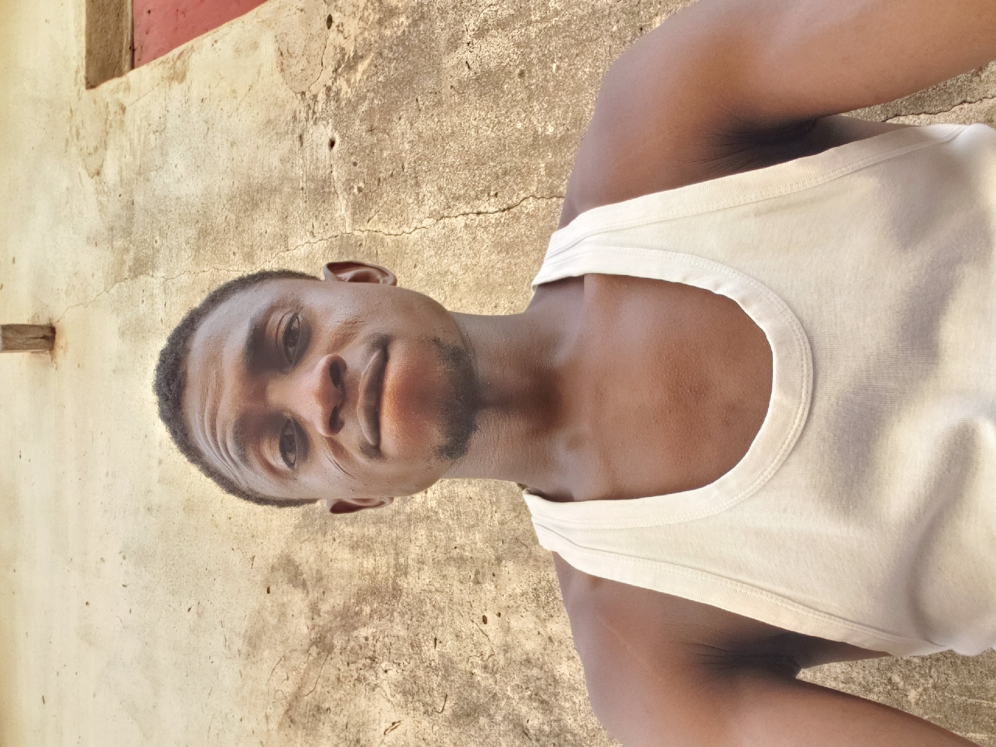 A head-and-shoulders portrait of a young Black man, likely a selfie, is captured against an outdoor, textured wall. The man, appearing to be in his late teens or early twenties, has dark brown skin, short dark curly hair, and light stubble on his chin and upper lip. He is wearing a plain white tank top. He looks directly into the camera with a neutral and calm expression.
The background consists of an old, light beige or pale brown concrete or stucco wall, characterized by numerous cracks and an uneven texture, suggesting age and exposure to the elements. In the upper left background, a dark wooden plank or beam is visible, while in the upper right, a section of a reddish-brown structure, possibly a window frame or door, can be seen.
The scene is brightly lit by natural daylight, indicating an outdoor setting during fair, possibly sunny, weather. There are no strong shadows, suggesting the sun might be high in the sky. The overall image gives a candid glimpse into a daily moment. The location is Adankolo, Nigeria. No visible text is present in the image. The image is oriented sideways, with the man's head facing the left side of the frame.