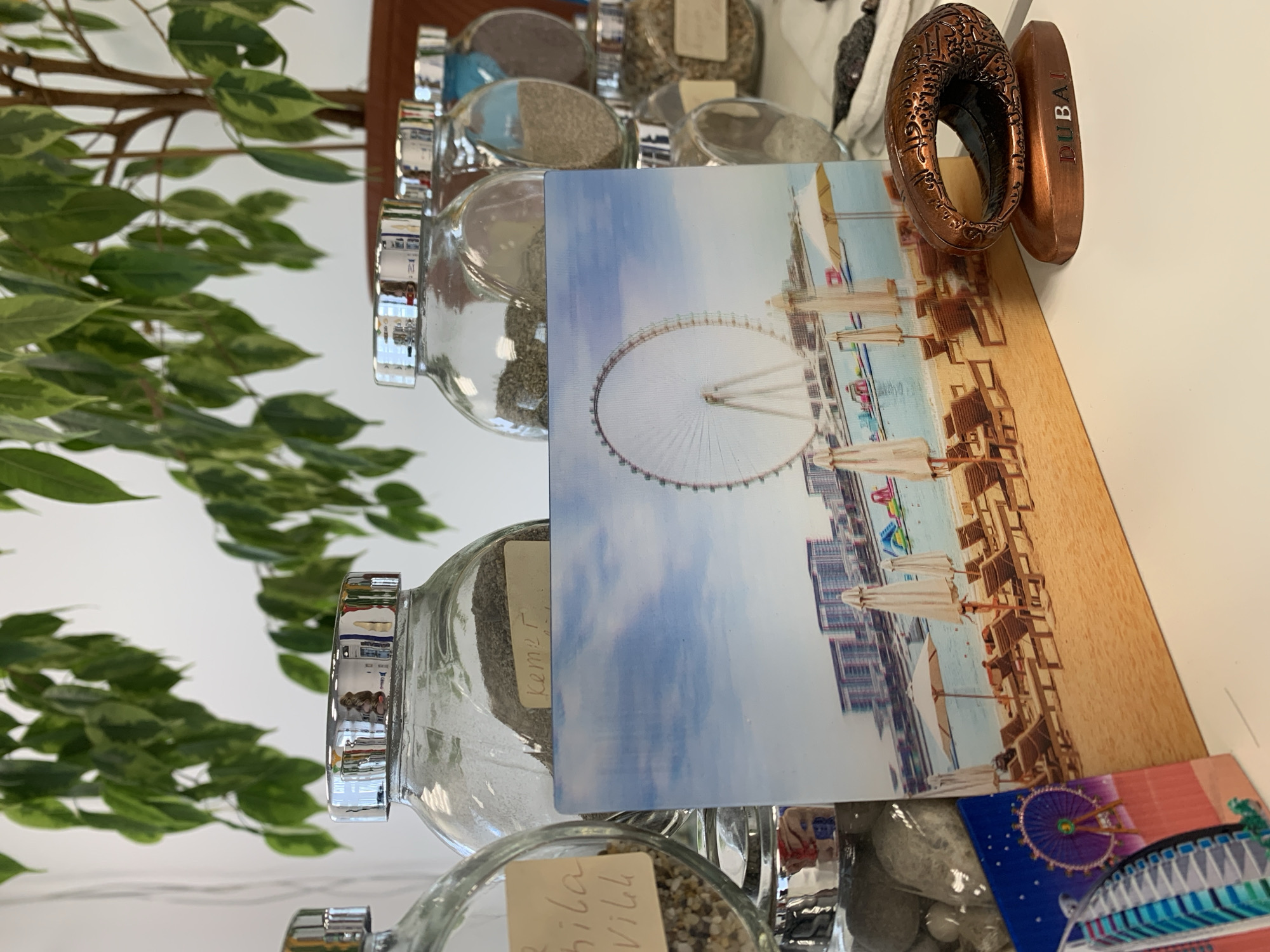 The image depicts a close-up, eye-level view of a white surface, likely a table, upon which several objects are arranged. In the foreground, a small, bronze-colored souvenir sits on a base that reads "DUBAI." The souvenir is an oval shape with intricate etched patterns and Arabic calligraphy. To the left of the souvenir, a rectangular postcard is propped up, displaying a photograph of a beach scene with lounge chairs, umbrellas, and a large Ferris wheel in the background, also featuring the text "DUBAI."
Behind the postcard, several glass jars are visible, arranged in a row. These jars have silver lids and contain various granular substances, possibly sand or small stones. One jar on the far left has a handwritten label with illegible text. Another jar in the center has a label that reads "kemer."
In the background, a large green leafy plant is prominent, its branches extending across the top of the frame. The wall behind the plant is white. The overall impression is that of a collection of travel souvenirs and natural elements displayed on a flat surface in an indoor setting. The location is identified as the city of Kommunar, Russia.