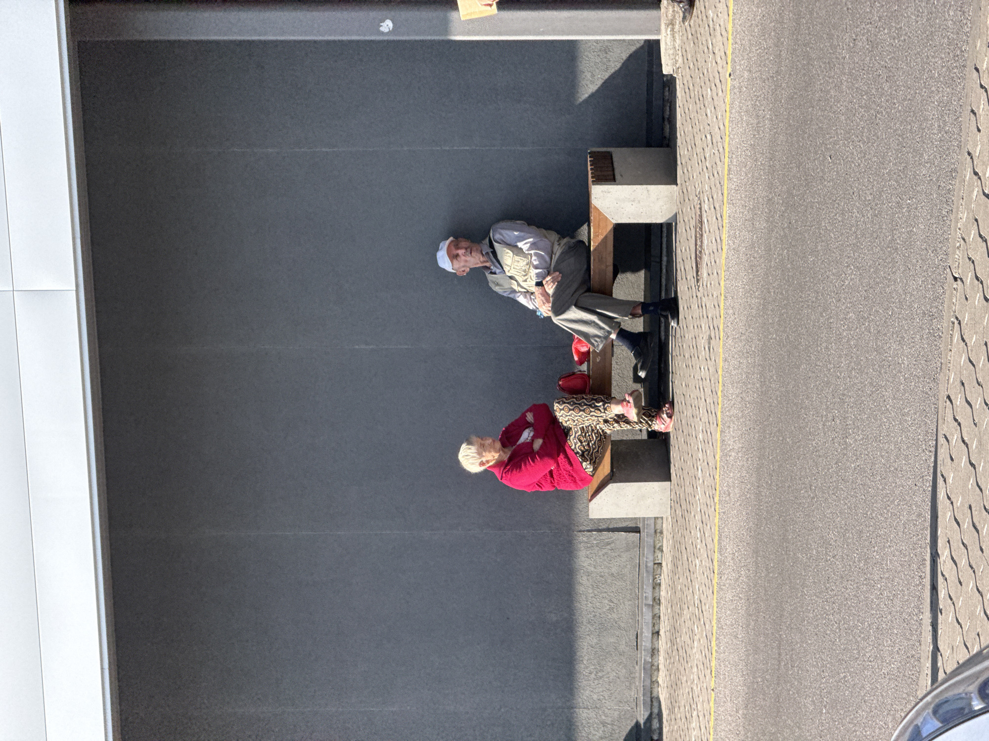 Two individuals are seated on a concrete and wood bench in front of a dark gray, textured wall. The woman on the left wears a bright pink knitted sweater, a white undershirt, and patterned trousers with leopard-print accents. Her arms are crossed, and she is looking downwards. A red handbag is placed on the bench next to her. The man on the right wears a light-colored vest over a long-sleeved shirt, gray trousers, a white cap, and dark shoes with dark socks. He is looking to his right. The bench is positioned on a light gray paved area, separated from an asphalt road by a thin yellow line. Sunlight casts a shadow from the wall onto the paved area behind the bench, indicating it is likely daytime. This scene appears to be a public space, possibly a bus stop or waiting area, within the city of Tallinn, Estonia.
