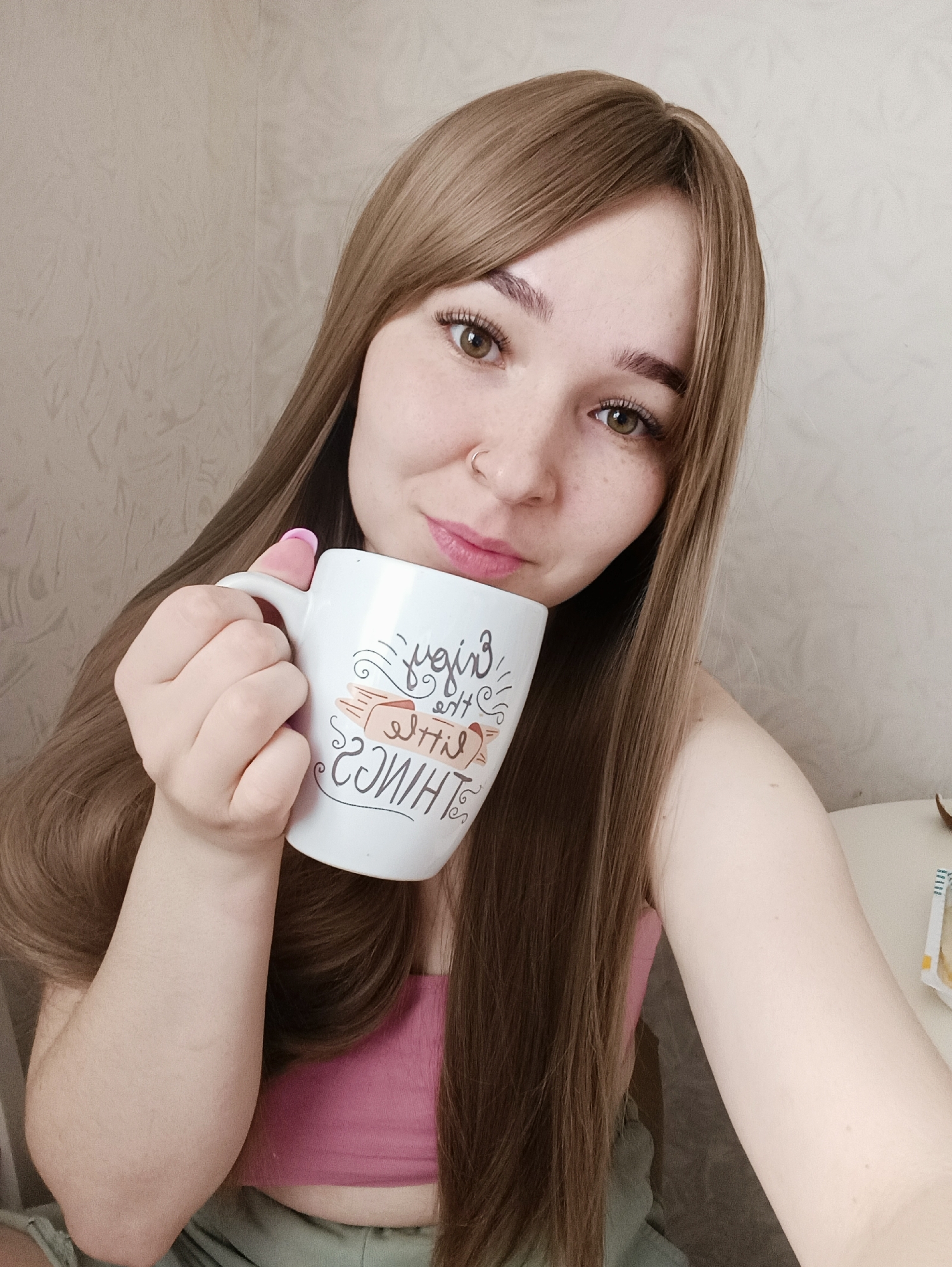 Location: Izhevsk, Russia.
A single female subject is depicted in a close-up, indoor setting. The female has long, straight, light brown hair parted slightly off-center and brown eyes. She has freckles on her nose and cheeks and a silver hoop nose ring in her left nostril. Her fingernails are painted light pink. She is wearing a pink strapless garment and what appears to be light green fabric below. The subject is holding a white ceramic mug with her right hand, positioned near her mouth. The mug features a dark-colored inscription, mirror-imaged, which reads "enjoy the little things." The background consists of a light-colored, textured wall. A light-colored, flat surface is visible on the right side of the frame. The subject is looking directly at the camera.