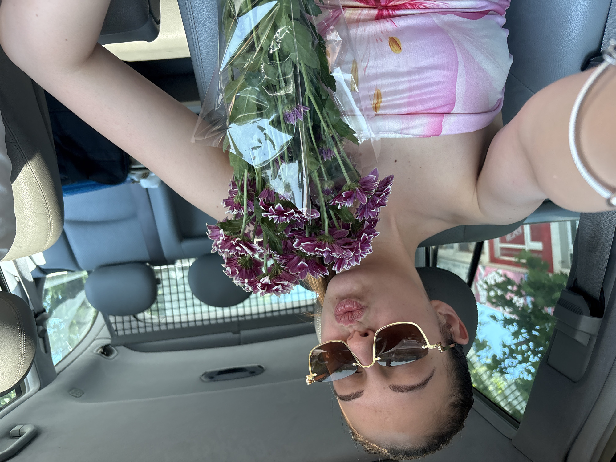 A female individual is depicted taking a selfie while seated in the driver's seat of a vehicle, in what is identified as Tsarevo, Bulgaria. She has light brown hair pulled back from her face and is wearing gold-framed, rimless sunglasses with dark brown lenses. Her lips are pursed in a pouting expression. She is wearing a strapless top featuring a pink and white floral or abstract pattern. Her left arm is raised, holding the camera, and a silver-colored bracelet is visible on her wrist. She is holding a bouquet of purple and white chrysanthemums, wrapped in clear plastic, in front of her torso.
The car's interior is visible, featuring grey upholstery on the seats (driver's seat and rear seats) and matching grey trim on the door panel and ceiling. Behind the rear seats, a metal grid or cargo barrier is present. Through the driver's side window, green foliage from trees and a blue sky are visible, along with a blurred background showing a building with red and white elements. The overall lighting suggests a bright, sunny outdoor environment.