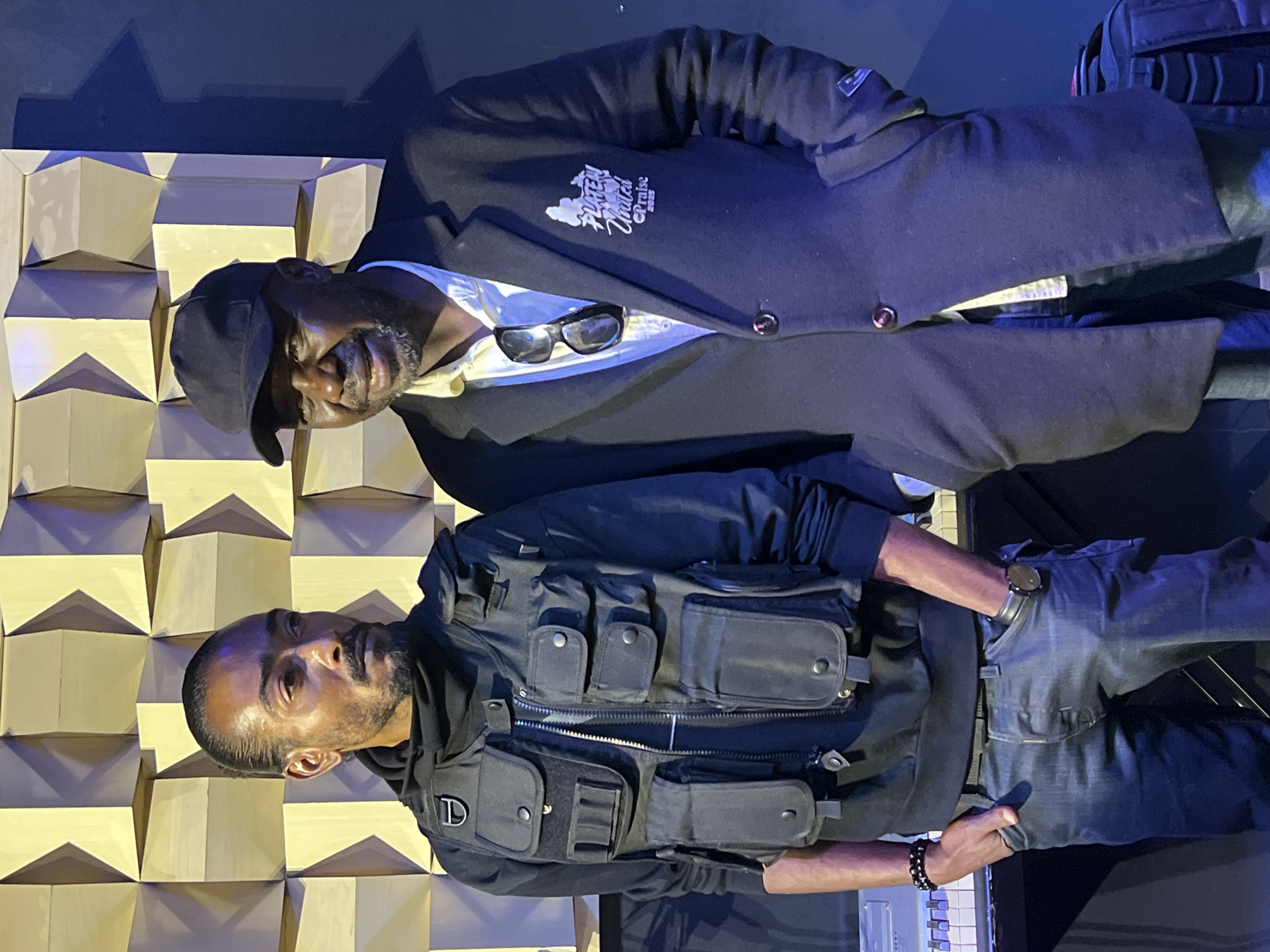 Two men are standing in what appears to be a recording studio. The man on the left has dark skin, a goatee, and is wearing a black hooded sweatshirt, a black tactical vest with multiple pockets, and dark blue jeans. He has a beaded bracelet on his left wrist and a watch on his right. The man on the right has dark skin, is wearing a black cap, a light-colored collared shirt, a dark blazer with a white logo on the left breast, and dark blue jeans. He has sunglasses clipped to his shirt. A keyboard is visible in the foreground on the left. The background features a geometric wall paneling. The lighting is dim with blue accents.