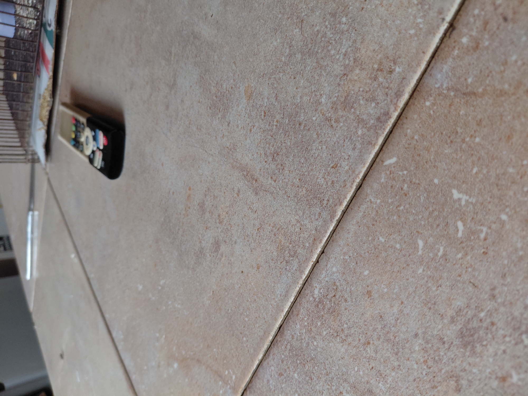 The image presents an oblique, overhead view of a tiled surface, likely a floor, located in Santa Ana, Venezuela. The tiles are light brown to beige in color, featuring a mottled, textured pattern resembling natural stone or aggregate. Visible grout lines run diagonally across the frame, clearly separating the individual tiles.
A black remote control, equipped with various colorful buttons (including red, blue, green, yellow, white, and grey), lies on the tiled surface in the upper left portion of the image. To its left, a section of a wire rack or cage structure is partially visible. Adjacent to the rack, there appears to be some crumpled material, possibly paper or fabric, displaying faint hints of red, white, and green. A subtle, vertical reflection, possibly from a nearby window or door frame, can be discerned on the far left edge of the frame. The scene is brightly and evenly lit, indicative of daytime. No readable text is visible on any of the objects.