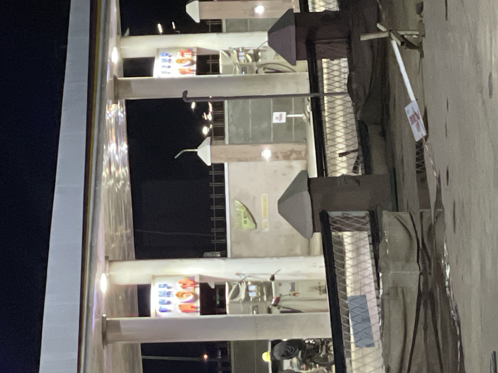 The image shows a partially lit gas station in Hayin Banki, Nigeria at night. Two gas pumps are visible under an awning. A sign displaying "FUEL" is illuminated above each pump. The station's exterior is a mix of concrete and metal grating, showing wear and some disrepair. A small sign with illegible text is affixed to a post near the pumps. The surrounding environment is dark and appears sparsely populated.