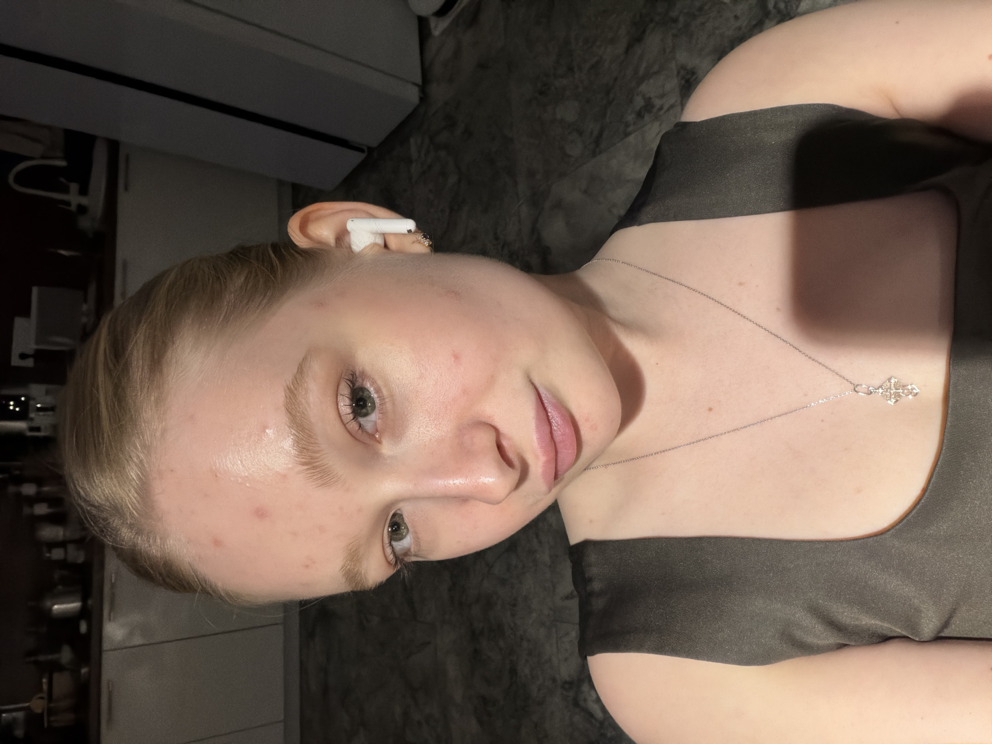A young, light-skinned woman with blond hair is shown from the shoulders up. She is wearing a dark top and a silver necklace with a cross pendant. She has an earbud in her left ear. The background is blurry but shows a dark surface and a white appliance. The location is unknown and there is no information to indicate it is in Rossiyskiy, Russia.