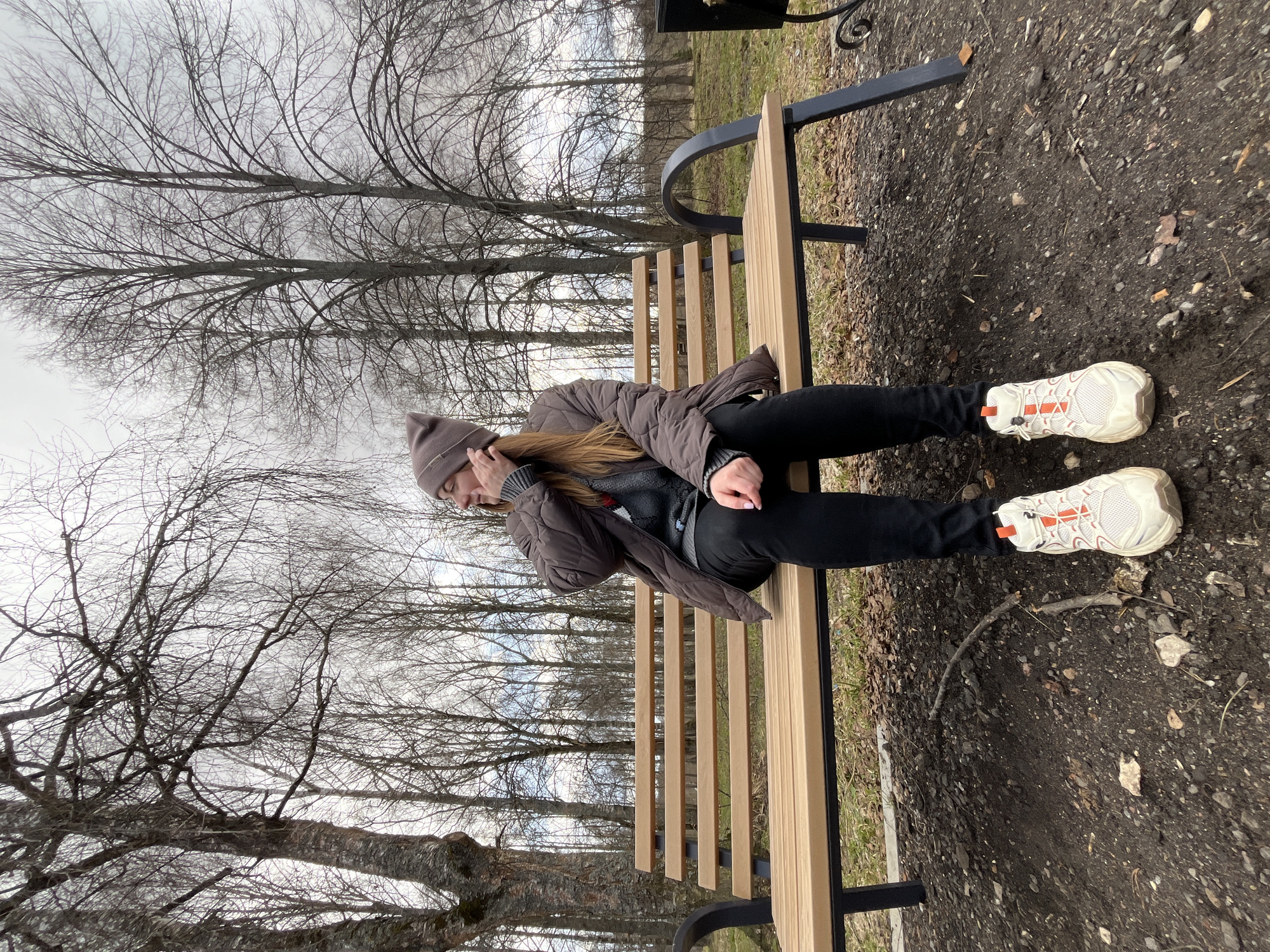 A young woman sits on a wooden bench in a park in Vysokovsk, Russia. She wears a brown quilted jacket, a grey sweater, black pants, and white sneakers with orange accents. A brown beanie covers her head. The park has bare trees, indicating a cold season. The ground is covered with dirt and small stones.