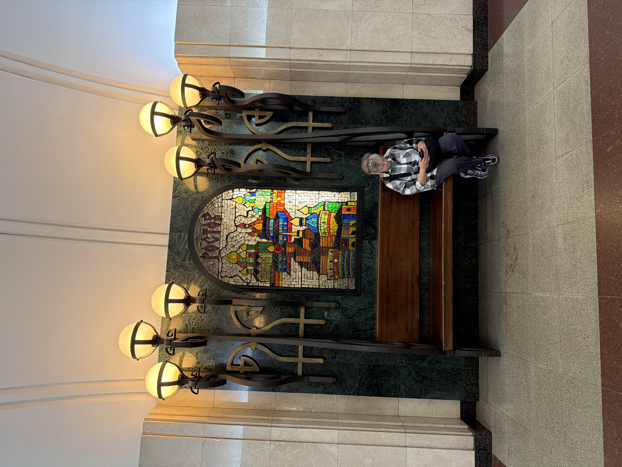 A woman sits on a wooden bench in a subway station, looking at her phone. She is wearing a black and white plaid shirt, dark pants, and athletic shoes. Behind her is a mosaic depicting a city with colorful buildings and domes, with the word "РОСТОВ" (Rostov) above it. The mosaic is framed by ornate metal sconces with globe lights on either side. The station features marble-clad walls and a tiled floor. This appears to be the Rostov station of the Moscow Metro, which is decorated with mosaics of Russian cities.