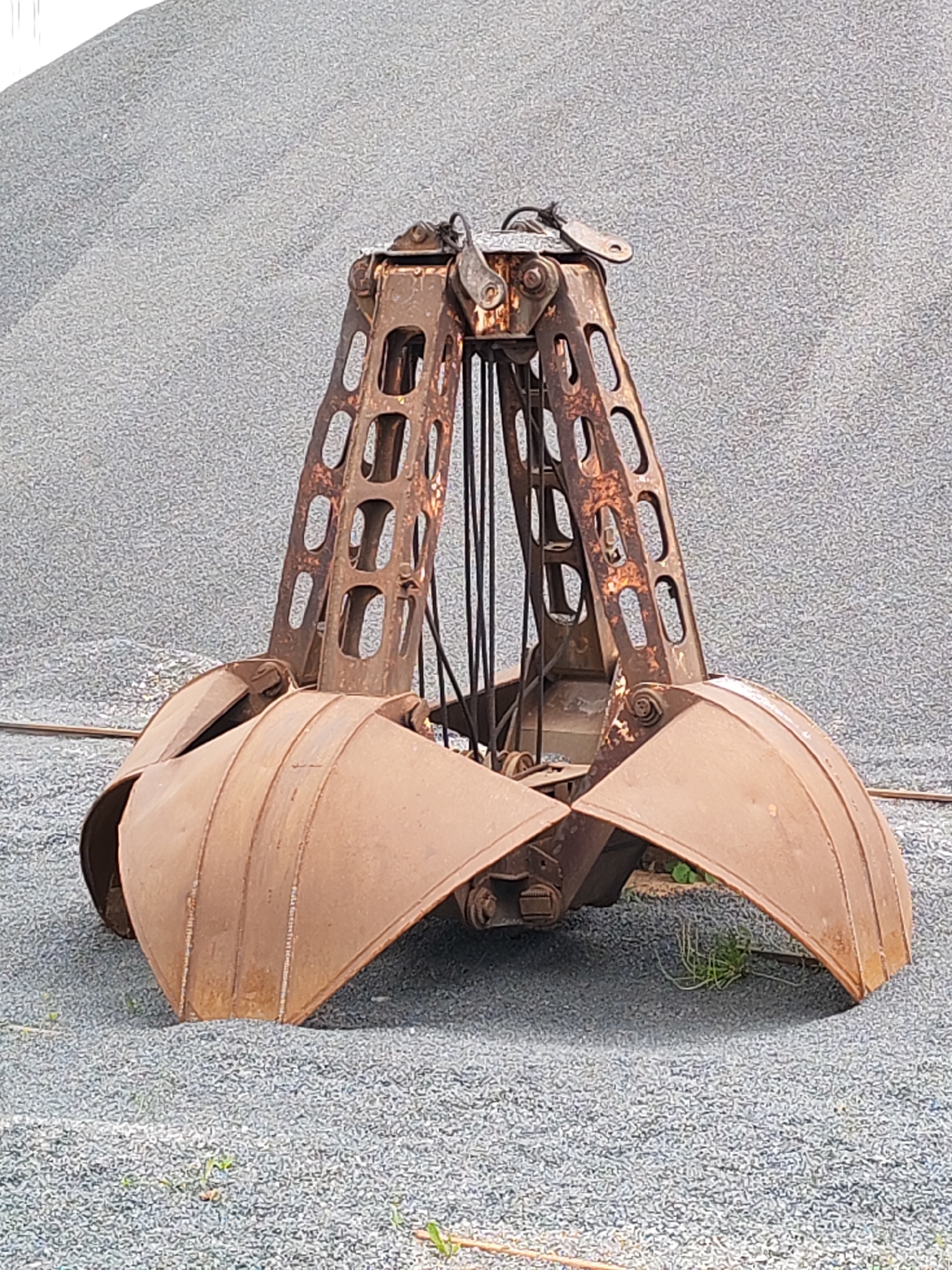 A large, rusty metal clam shell grab, possibly for a crane, sits on a gravel surface. The grab has an A-frame structure with rectangular cutouts and cables running down to the curved, open clamshell buckets. The buckets are also rusty. In the background is a large pile of gravel. There is vegetation visible near the bottom of the image. The exact location in Rybinsk, Russia is not apparent from the image. There are no people in the image.