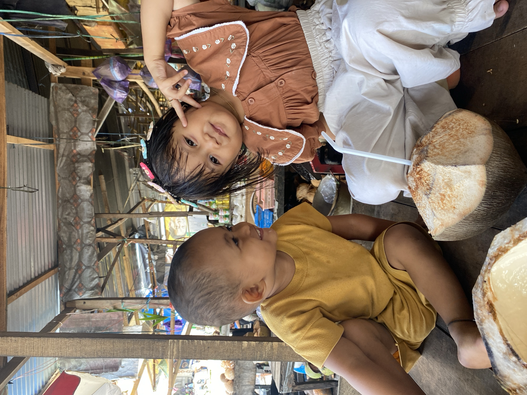 In Ta', Indonesia, two young children are seated on a wooden floor, likely within a market stall or open-air structure. The child on the left, wearing a yellow t-shirt, looks to their right and smiles. The child on the right, adorned with colorful hair clips, wears a brown blouse and cream-colored pants. This child poses, making a hand gesture near their face, and looks towards the camera. In the foreground, two halved coconuts are visible, one with a straw inserted, suggesting they are enjoying the coconut water. The background reveals the rustic construction of the stall, with exposed wooden beams, corrugated metal roofing, and various items hanging, indicative of a marketplace environment.