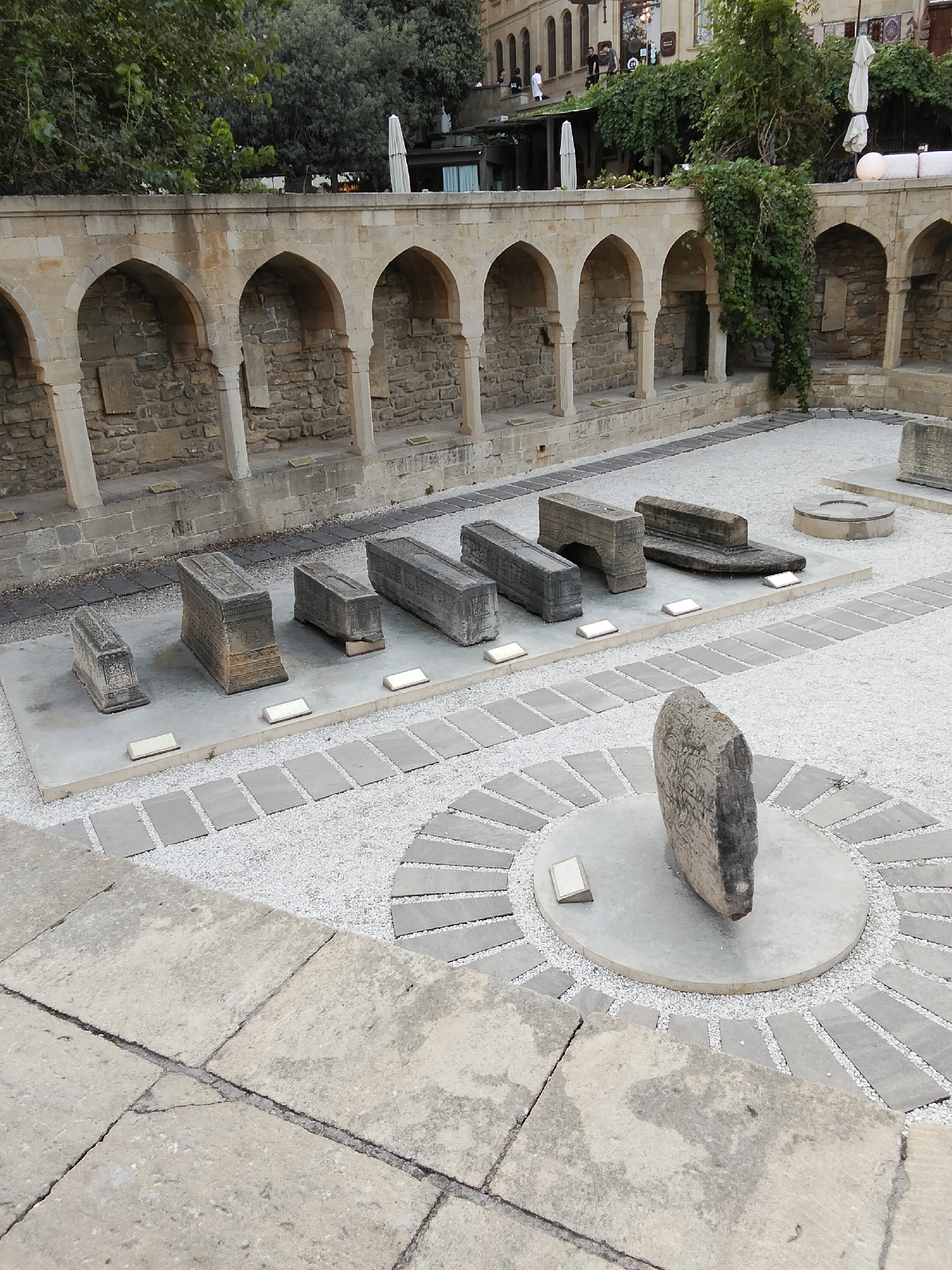 The image depicts an archaeological site in Sabail Raion, Azerbaijan, likely the Shirvanshahs' Palace complex or a related historical area. In the foreground, large, worn stone slabs form a courtyard. Leading into the middle ground is a pathway composed of smaller, evenly spaced grey rectangular stones interspersed with fine white gravel. This pathway leads to a circular display area featuring a tall, weathered grey stone monument inscribed with intricate patterns, standing on a concrete pedestal. Surrounding this central monument, more grey rectangular stones are arranged in a radial pattern within a bed of white gravel.
Further back, a series of stone sarcophagi and ancient stone artifacts are displayed on concrete platforms. These objects are rectangular and appear to be carved from dark grey stone, some with visible inscriptions or decorative patterns. They are arranged in rows, with small white rectangular markers placed in front of some.
Behind the sarcophagi, a long, arched stone structure made of weathered, tan-colored stone forms a backdrop. This structure features multiple arched alcoves with stone walls and pillars. Ivy and other greenery cascade from the top of this structure and from other walls in the background, suggesting an outdoor setting with historical architecture. In the distance, buildings with tan facades and arched windows are visible, indicating an urban environment beyond the archaeological site. Umbrellas and some people can be seen on a higher level behind the stone structure, suggesting the presence of a cafe or restaurant. The overall scene is one of historical preservation and display within a city context.