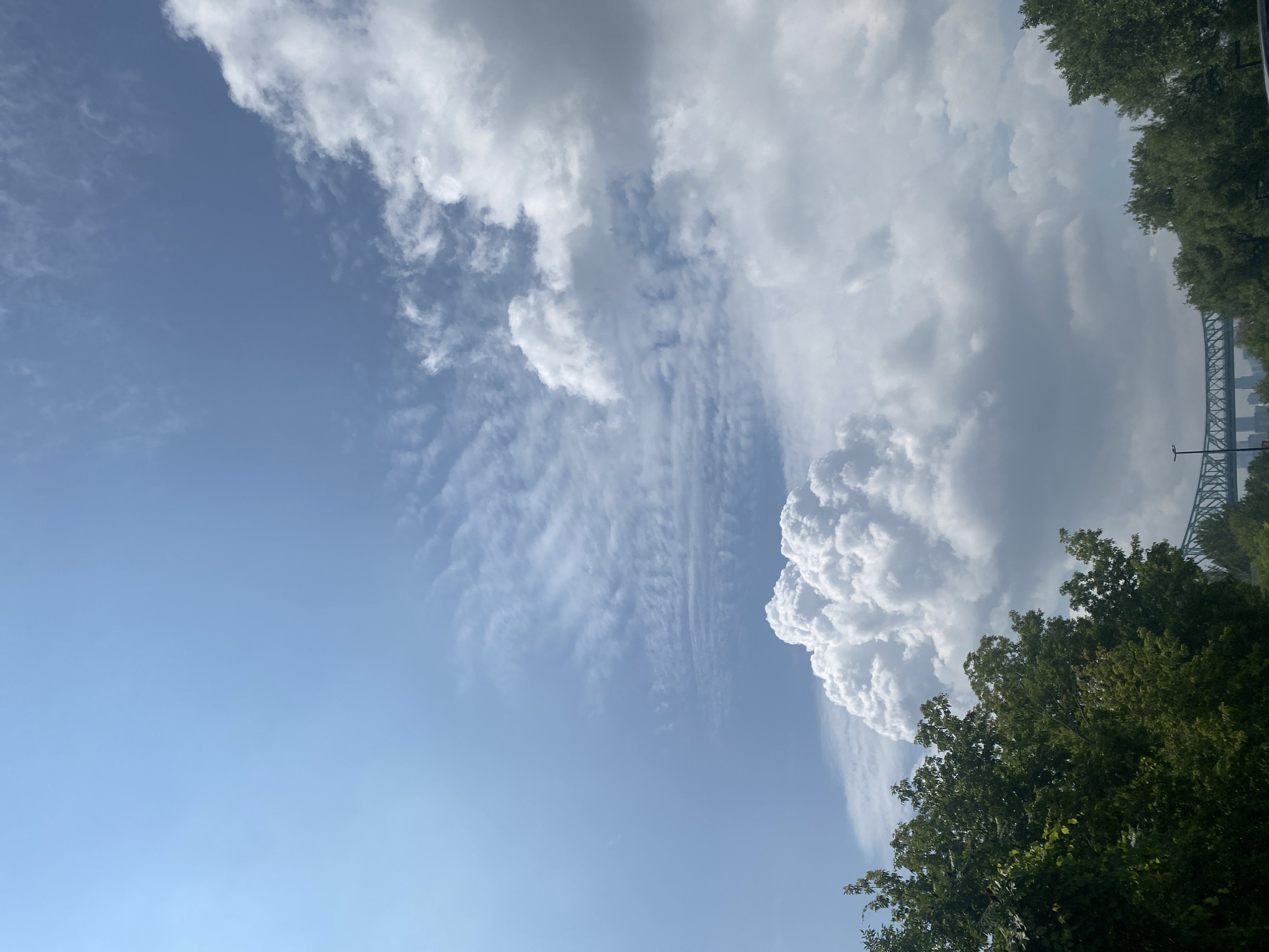The image captures a view of the sky over Montreal, Canada, with a prominent cloud formation dominating the center. The sky is a clear blue in the upper left, transitioning to wispy cirrus clouds and then to larger cumulus clouds on the right. In the foreground, lush green trees line the bottom of the frame. In the distance, a bridge with a green truss structure is visible, suggesting an urban setting. The overall scene suggests a bright, partly cloudy day.