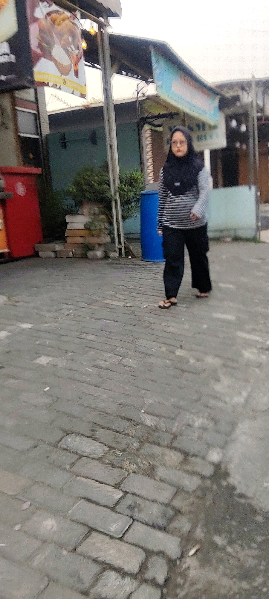 A full-body, slightly blurry shot captures a woman walking on a cobblestone street in front of a building. She is wearing a black hijab, glasses, a striped long-sleeved shirt, and black pants. Her feet are bare, shod in flip-flops. To her left, a red trash can stands next to a stack of bricks and a small bush. A large blue barrel is visible behind her, to the right of the building's teal-colored wall. A sign depicting food is displayed on the left side of the building, and another sign, mostly obscured by the woman, hangs above her on the right. The overall lighting suggests it is daytime, likely overcast, in Mekarmukti, Indonesia.
