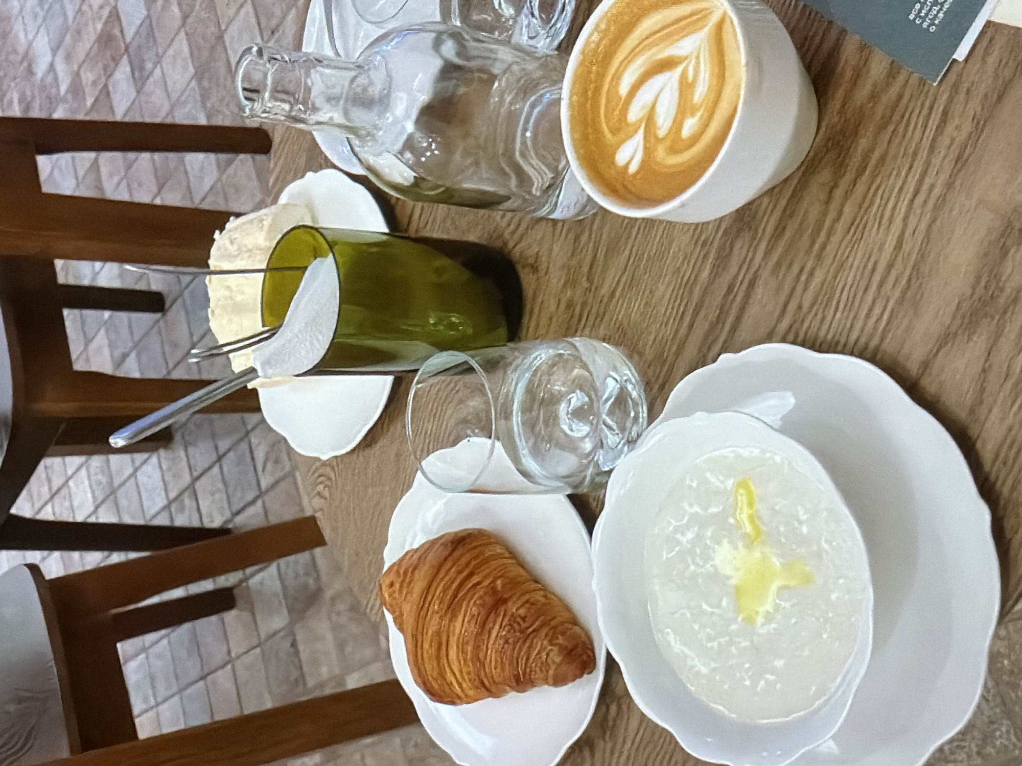 The image depicts a table setting in Unknown, Unknown, with various items arranged on a wooden surface.
On the table, there is a cup of coffee with latte art, a clear glass bottle, a piece of cheese with silverware on a plate, a green object, a glass, a croissant on a plate, and a bowl of white porridge with a yellow topping. The table is adjacent to a wooden chair, which is partially visible. The background features a tiled floor.
