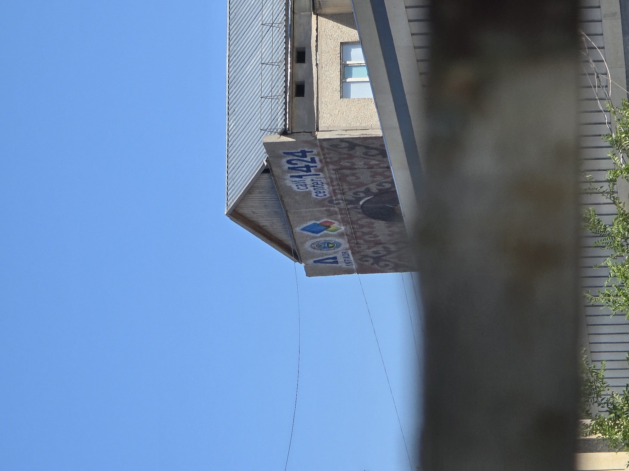 A building with a metal roof and light-colored siding is visible against a clear blue sky. A large mural covers a section of the building's facade, featuring a blue "A" symbol with colorful shapes and the words "call center 1424" in blue lettering. Below the text, a pattern of red and brown abstract designs adorns the wall. A window with white panes is visible on the right side of the building, and two small dark rectangular openings are located above it. A wire stretches horizontally across the sky. The foreground is obscured by a blurred dark object, possibly a fence or barrier, with greenery visible below it. The image appears to be taken in Qaratau, Kazakhstan.