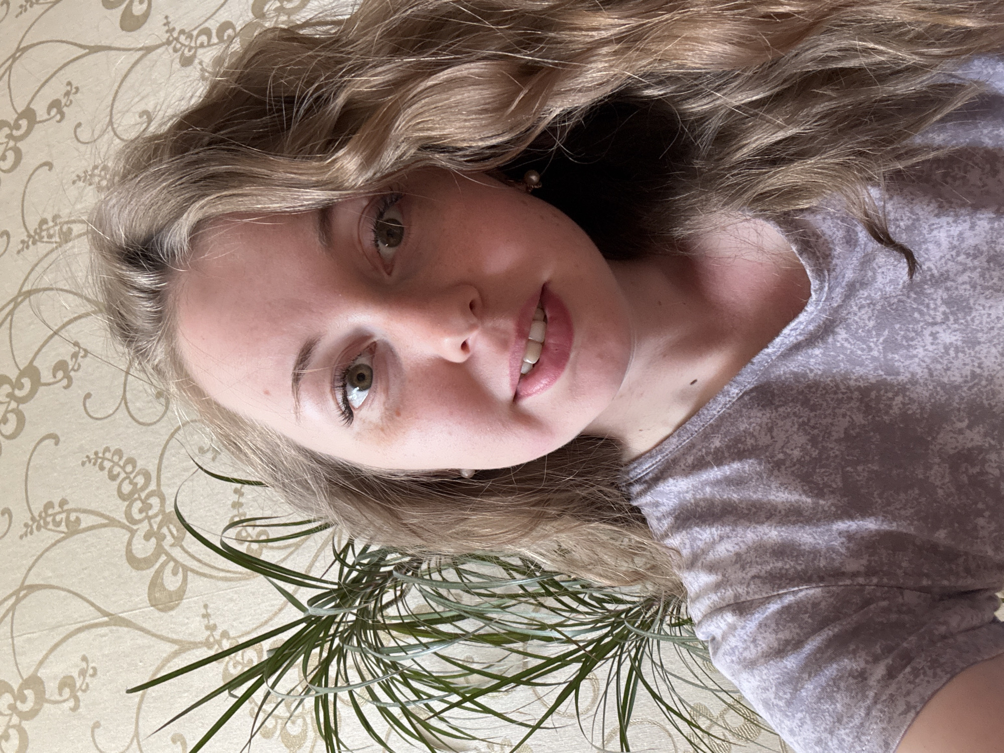A Caucasian female with light brown, curly hair and fair skin is pictured in a selfie. She has green eyes and is wearing a grey t-shirt. The background includes a beige patterned surface and green plant fronds. The location is Unknown, Unknown.