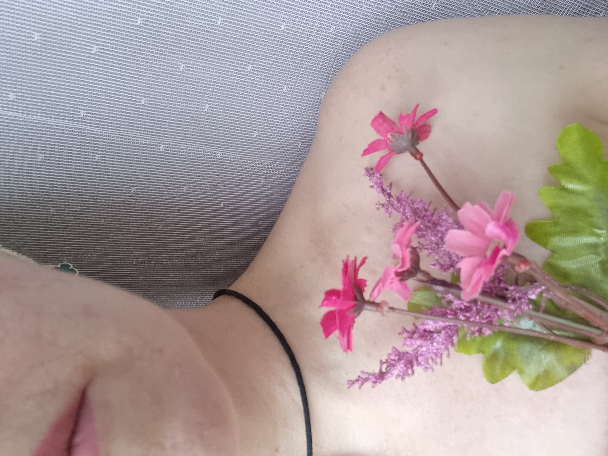 A close-up shot depicts a person, likely a woman, lying down. Pink flowers are placed on her shoulder, while a thin, black necklace adorns her neck. The background features a textured, gray surface. The image was taken from a high-angle perspective. The location is not discernible from the image.