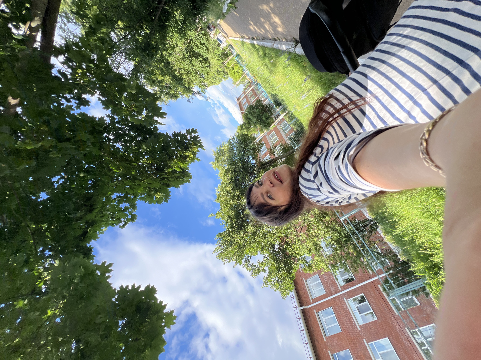 This is a selfie taken outdoors with a low-angle perspective, looking up towards the sky and surrounding environment. The main subject is a young woman with long, dark hair and bangs, wearing a blue and white striped t-shirt and a gold bracelet. She is looking directly at the camera with a neutral expression.
The setting appears to be a residential area, possibly a university campus or apartment complex, in Noviy, Russia, based on the context provided. In the background, there are several multi-story brick buildings with visible windows. A well-maintained lawn and a paved walkway are also present. Lush green trees with dense foliage frame the scene, with their leaves and branches reaching towards the sky. The sky is a bright blue with scattered white clouds, suggesting it is daytime with good weather, likely sunny. Sunlight filters through the trees, casting dappled shadows on the ground and on the woman's clothing. A black object, possibly a stroller or a bag, is visible near the woman. The overall mood is relaxed and casual, indicative of a sunny day spent outdoors. No visible text is present in the image.