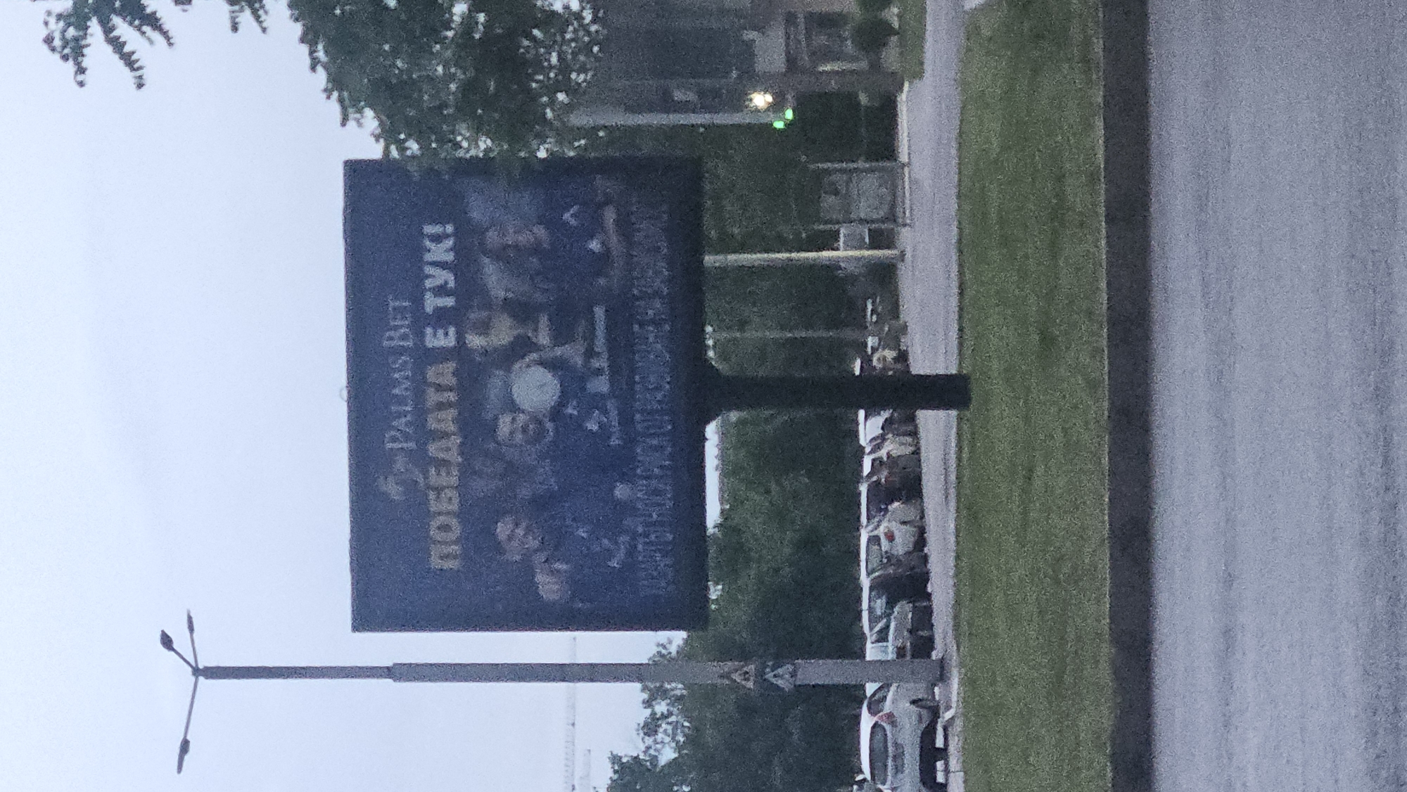 The image depicts an outdoor scene in Plovdiv, Bulgaria. A "Palms Bet" advertising billboard is prominently featured, displaying the text "ПОБEДATA E TУK!" (Victory is Here!). The billboard is positioned near a street with parked cars and a grassy area. A street lamp is visible in the foreground. The weather appears overcast.