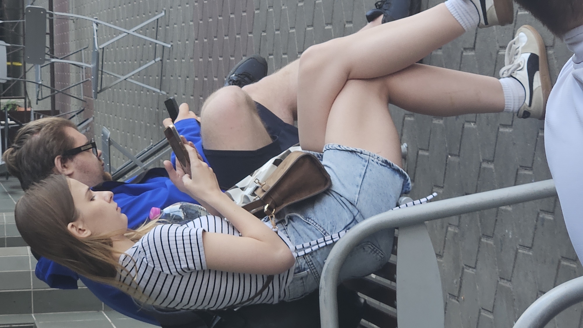 A young woman with blonde hair and a striped shirt sits on a bench next to a man with brown hair and glasses. She is looking at her phone, which she holds in her hands. The man is also looking at his phone. They are both sitting outdoors on a paved area with some chairs in the background. The image appears to have been taken in the city of Kysak, Slovakia.