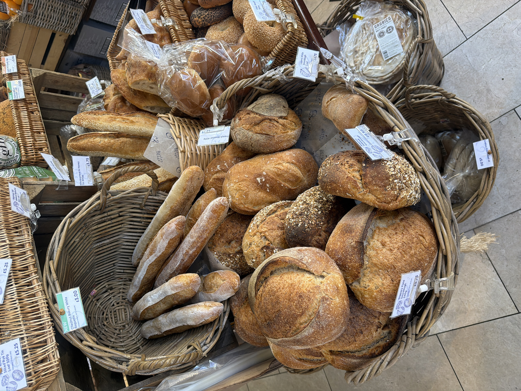 This image shows an assortment of baked goods displayed in baskets at a bakery or market. The location is most likely in the city of London, United Kingdom, as evidenced by the presence of price tags in pounds sterling. The main subjects are various types of bread, including baguettes, loaves, and rolls, arranged in woven baskets. Several price tags are visible, indicating the name of the bread and the corresponding price (e.g., "Chestnut Bakery White Baguette £3.00," "French Bloomer 400g £2.75"). The environment appears to be an indoor market or shop setting. The floor is tiled, and there are other baskets and products visible in the background, suggesting a commercial food setting. There are no people visible in the frame, but the presence of the bread and the signage indicates a retail operation.