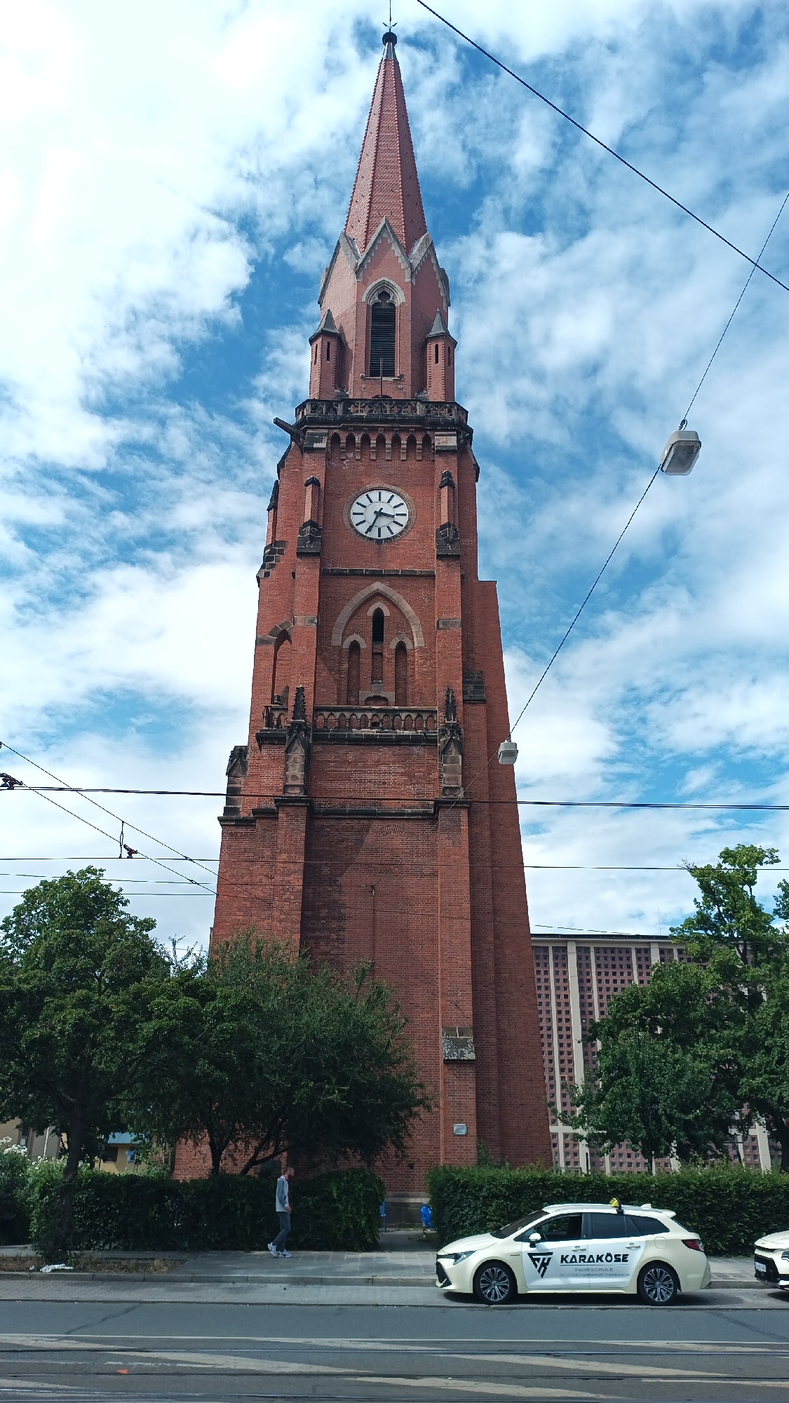 The image captures a street scene in Nuremberg, Germany, with a prominent red brick church tower dominating the center. The tower features a pointed steeple, a clock face showing approximately 10:20, and gothic architectural elements. In the foreground, a white taxi with the branding "KARAKÖSE" is parked on the street. A person in a grey sweatshirt and dark pants is walking on the sidewalk in front of a hedge and trees. Overhead, power lines and streetlights are visible against a blue sky with scattered white clouds. In the background, a multi-story building with a grid-like facade can be seen.