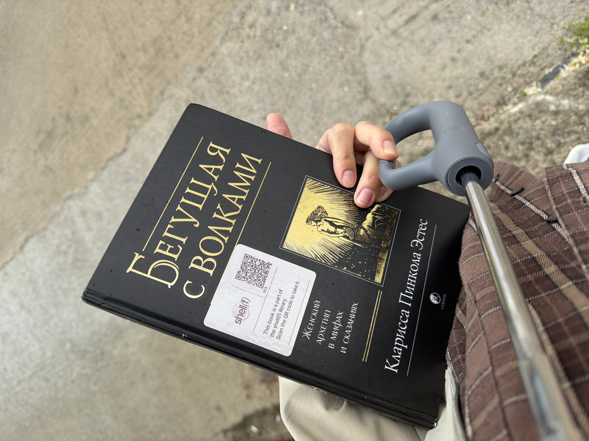 A close-up, high-angle outdoor shot captures a person's right hand holding a book in Budva, Montenegro. The book has a black hardcover with gold Cyrillic lettering and a golden illustration depicting a figure and a wolf. The title visible is "БЕГУЩАЯ С ВОЛКАМИ" (Women Who Run With the Wolves) and the author is "Кларисса Пинкола Эстес" (Clarissa Pinkola Estes). Below the illustration, the subtitle reads "ЖЕНСКИЙ АРХЕТИП в мифах И СКАЗАНИЯХ" (Female Archetype in Myths and Tales), published by "СОФИЯ" (Sofiya).
A white sticker on the book cover displays a QR code, the text "shell(f)", "323", and "This book is a part of the shell(f) library. Scan the QR code to take it." The hand is also seen gripping a gray, ergonomic handle with a silver metal pole extending from it, possibly from a stroller, cane, or trolley. Part of a brown and tan plaid fabric, likely clothing or a bag, is visible to the right. The background consists of a light-colored, textured concrete or paved ground, suggesting an outdoor or sidewalk setting during daytime. The image depicts a moment of carrying a book, potentially while in transit or outdoors.