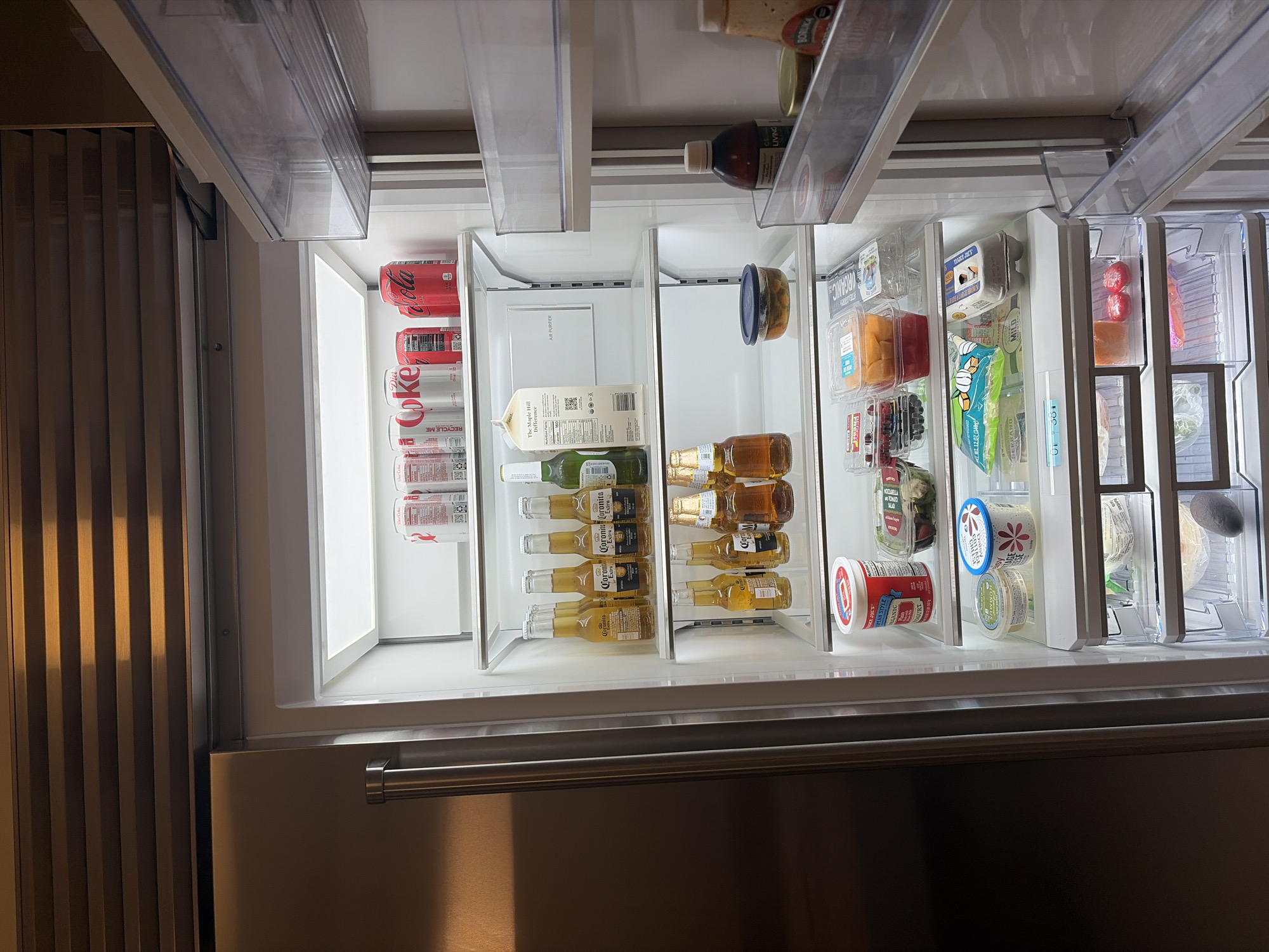 Detailed analysis of the open refrigerator reveals a variety of food and beverage items neatly arranged on the shelves. The leftmost shelf contains numerous cans of Diet Coke. Adjacent to this shelf is a selection of items including a carton of Maple Hill Organic milk, several bottles of Corona Extra beer, and a mix of clear and amber-colored bottles. The rightmost shelves display assorted containers with yogurt, cheese, fruits, and salads. Above the shelves are additional containers and bottles.
The location is not specified or indicated by the visual information, and therefore any claims of it being in the city of Key Biscayne, United States of America or any other location is not supported by the provided image.