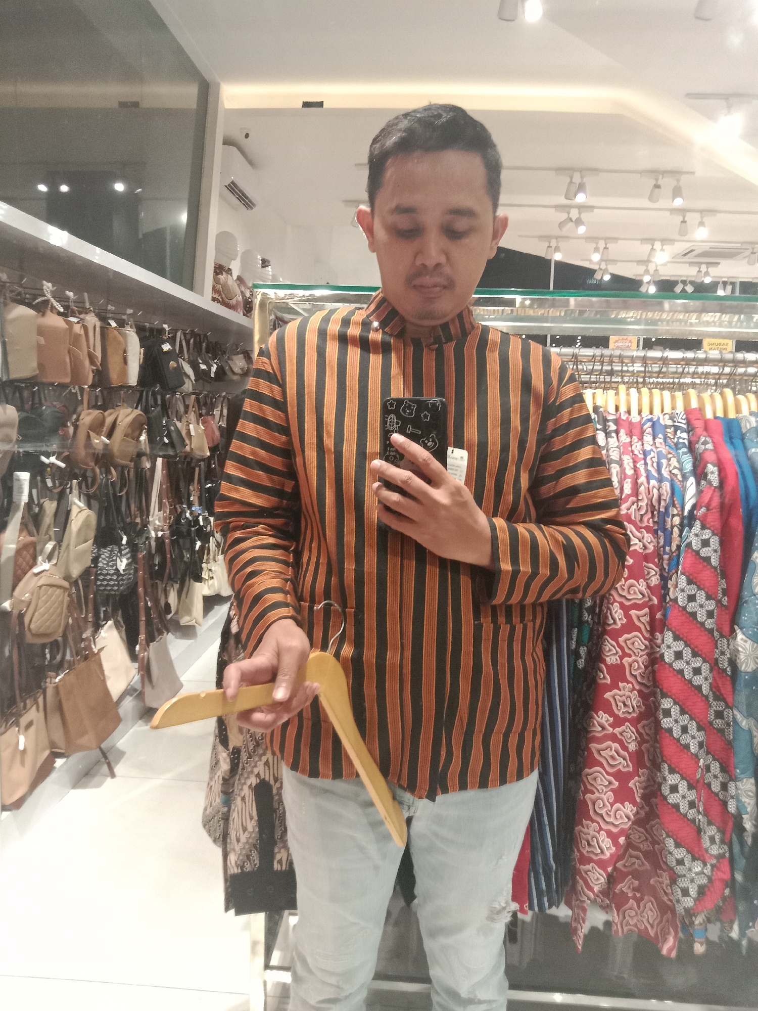 A man stands in what appears to be a retail store in Semarang, Indonesia, taking a selfie. He is wearing a striped long-sleeved shirt in orange and black, and light-colored ripped jeans. In his right hand, he holds a smartphone with a black case featuring cartoonish white characters. In his left hand, he holds a wooden clothes hanger. Behind him, shelves are stocked with various bags, and a rack displays colorful batik fabrics. The store is well-lit with overhead lighting, and mirrors reflect the merchandise and ceiling. The overall impression is of a casual shopping environment.