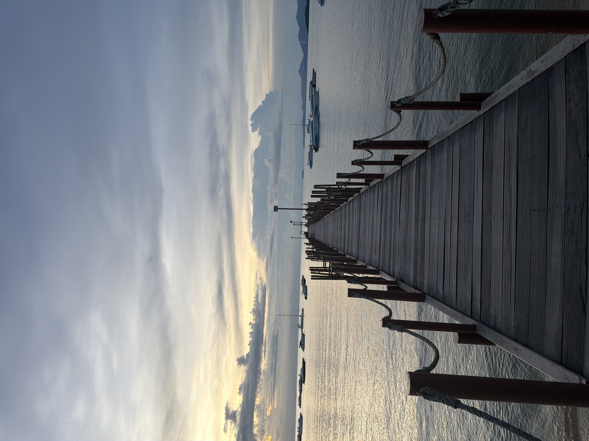 A long wooden pier extends into a calm body of water under a cloudy sky. The pier has weathered wooden planks and is lined with reddish-brown posts with ropes strung between them. Several boats, including speedboats and a sailboat, are anchored on the water in the distance. The sky is a mix of gray clouds and streaks of yellow and orange light, suggesting either sunrise or sunset. The water is a muted blue-gray, reflecting the sky. The scene is located in Baan Bang Rak, Thailand, a coastal area known for its piers and boat activity. There are no people visible in the image.