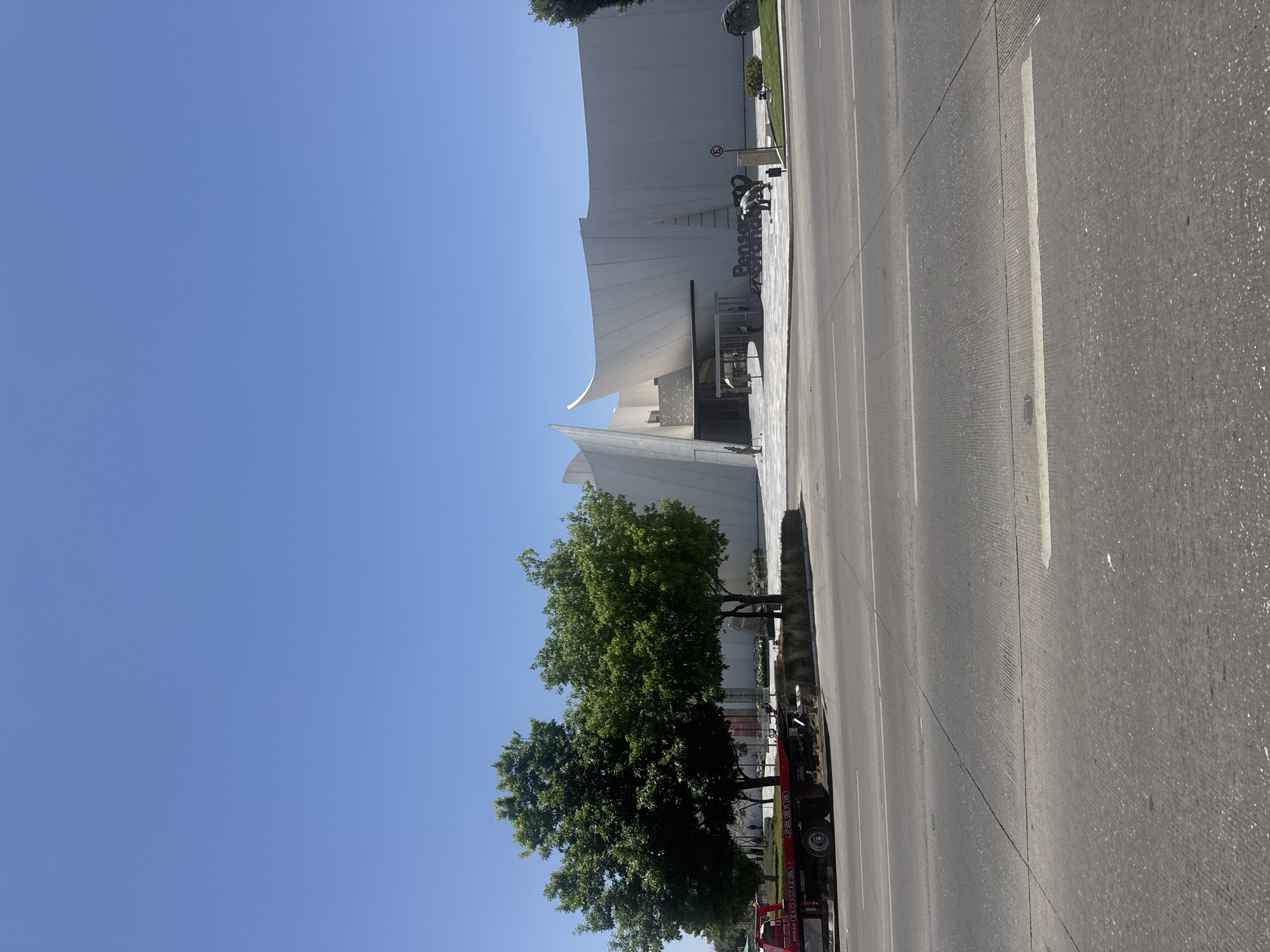 This image, taken in San Bernardino Tlaxcalancingo, Mexico, presents a bright, sunny daytime street scene dominated by a striking modern building. The sky is a clear, cloudless blue, indicating pleasant weather conditions.
The main subject is a large, white building with a unique, contemporary architectural style. It features prominent curved lines and sharp angles, giving it a sculptural, almost futuristic appearance. In front of the building, on a paved plaza, several artistic elements are visible. A dark-colored sculpture or sign prominently displays the Spanish text "Piensa en Grande" (Think Big). Further back, another large, dark, spherical object is part of the landscaping.
A row of vibrant green trees stands between the building's plaza and the street, adding natural color and shade to the urban environment. To the right, a wide, multi-lane concrete road stretches into the distance, marked with clear white lines. A red circular sign with a diagonal line, possibly indicating a traffic restriction, is visible near the building's entrance. On the far right edge of the image, partially obscured by the trees, a red vehicle, possibly a construction lift or truck, shows the numbers "63313" and "88003" and a partial word "BLECO" on its side.
The scene depicts a static, everyday view; no specific event or activity is taking place, and no people are distinctly visible. The overall impression is one of modern urban design set against a clear, bright sky.