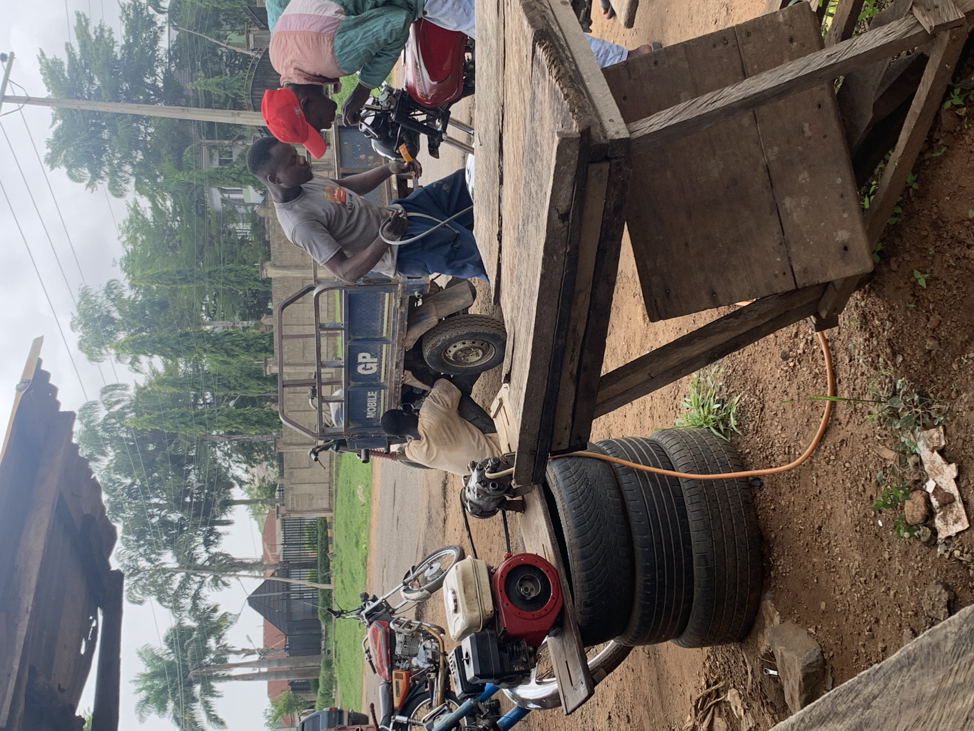 The image captures an outdoor scene, likely a roadside or open area, in Gbeleko, Nigeria, during the daytime with an overcast sky. The primary activity involves multiple men engaged in what appears to be vehicle maintenance or service.
In the foreground and midground, several men are visible. One man in a red cap and a light-colored top leans over a red motorcycle, interacting with another man in a grey t-shirt and blue trousers. The man in the grey t-shirt holds a white hose or cable, possibly connecting it to the tricycle or a piece of equipment. The motorcycle is positioned near a blue three-wheeled cargo tricycle. On the side of this tricycle, the words "MOBILE" and "GP" are visible.
Further back, another man, dressed in a light-colored long-sleeved shirt, is crouched down near the rear wheel of the blue tricycle, seemingly working on it or an engine assembly nearby. Adjacent to this area is a makeshift mobile tire repair setup, featuring a small red and white engine/generator, a pump or compressor, and a stack of three black tires on the dirt ground. An orange hose runs from this equipment.
To the right, a large, rustic wooden stall or platform dominates a portion of the frame. The ground is primarily reddish-brown dirt with patches of sparse grass and small plants. In the background, tall green trees, including palm trees, line the road, with some residential buildings faintly visible behind them. The overall impression is one of bustling roadside commerce or service in a developing area.