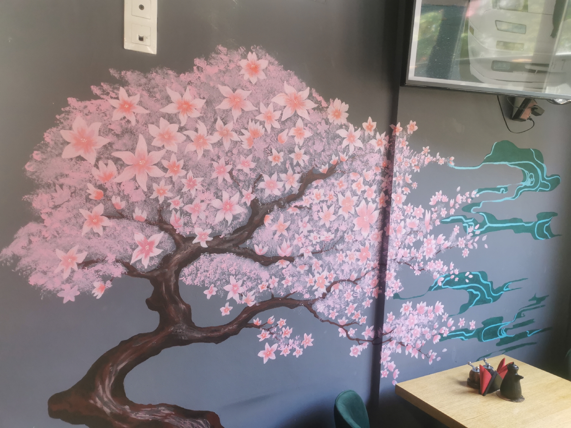 This media file captures an indoor scene in Blagoevgrad, Bulgaria, featuring a large mural dominating a grey wall, likely within a cafe or restaurant. The central subject of the mural is a vibrant cherry blossom (sakura) tree. Its thick, dark brown trunk and branches extend from the bottom left, spreading across the wall. The tree is densely covered with numerous pink blossoms, depicted with varying shades of pink and some individual flowers featuring lighter centers.
To the right of the tree, several stylized green and light blue cloud-like shapes are painted, complementing the East Asian aesthetic of the mural. In the bottom right corner of the image, a light-colored wooden table is visible. On the table, there's a condiment holder containing red napkins, a salt shaker, and a pepper shaker. Beside it sits a dark, possibly ceramic, figurine resembling a cat. A dark green chair back is partially seen to the left of the table.
In the upper left portion of the wall, a white electrical outlet with two switches or sockets is mounted. In the top right, a rectangular, framed object, possibly a television or a large picture, is fixed to the wall. This object reflects the front of a white vehicle, suggesting a window or proximity to an exterior space. A power adapter and cables hang below this reflective surface. No direct activity is taking place, but the setup indicates a space ready for dining. The overall atmosphere, conveyed by the soft pinks and calm grey, appears peaceful and inviting. There is no visible text in the image.