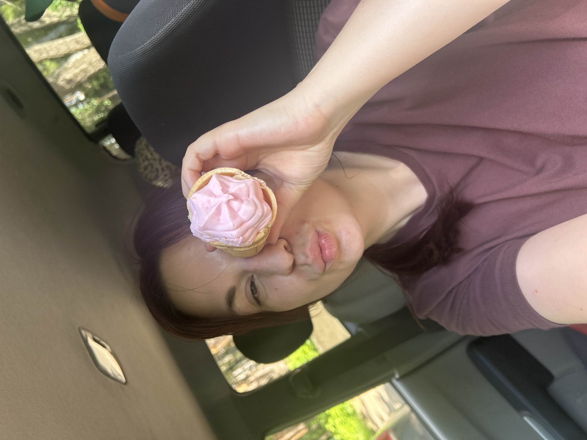 In a car, a woman with dark hair is holding a pink ice cream cone up to her right eye, puckering her lips. She is wearing a maroon t-shirt. The sun is shining, indicating it is daytime. Outside the car, trees and greenery are visible, suggesting a warm-weather location. The interior of the car is dark and textured, with the black seat of a car visible.