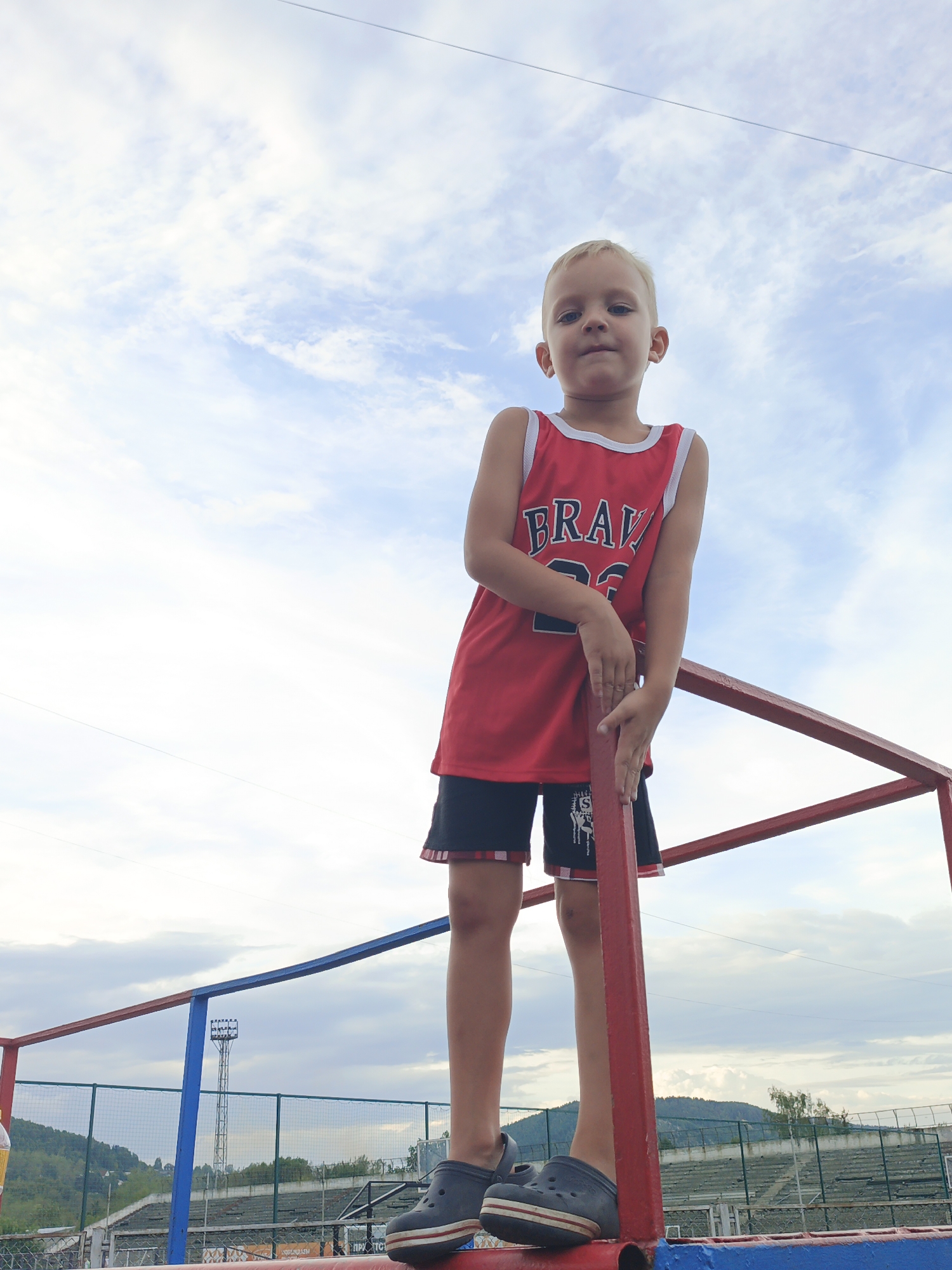In Zlatoust, Russia, a young boy with blond hair stands on a red metal railing against a cloudy sky. He is wearing a red tank top with "BRAVO" and "23" printed on it, black shorts with red trim, and dark gray Croc-style shoes. His hands are resting on the railing. In the background, a stadium with bleachers and a light tower is visible, surrounded by green hills. The scene appears to be outdoors, possibly at a sports facility.