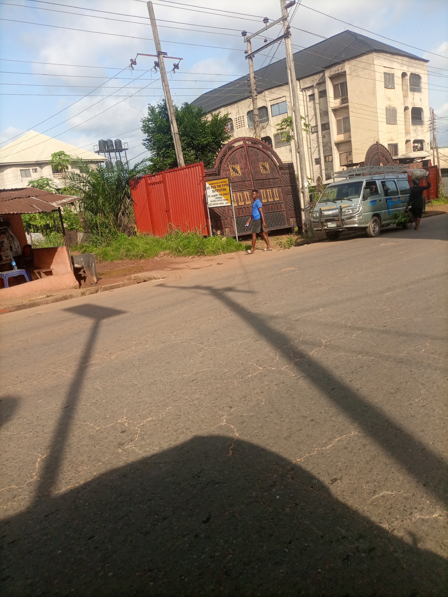 This image depicts a street scene in Nkpor, Nigeria, with a focus on a building and a gate. The building, appearing to be a multi-story structure, is light-colored with dark-tinted windows and an overall aged facade. In front of it is a large, ornate, dark metal gate with decorative elements, possibly marking an entrance to a property or institution. A red corrugated metal fence is situated to the left of the gate, bordering a grassy area.
A silver minivan is parked near the gate. To the left of the gate, a young man in a blue t-shirt and dark shorts is walking. Further to the left, a small roadside structure with a rusted metal roof is visible. The street itself is paved, with visible cracks and patches, suggesting wear and tear. Overhead, telephone poles and wires crisscross the sky, indicating the presence of utilities. The lighting suggests it is daytime, with shadows cast on the street. A woman carrying a load on her head is walking away from the minivan and towards the gate. The environment appears to be urban or semi-urban, with vegetation present around the buildings and along the roadside. The text on a yellow sign attached to the gate is partially visible and reads "ST. PIUS THE FIFTH OUTSTATION" and mentions "SUNDAY MASS - 7AM".