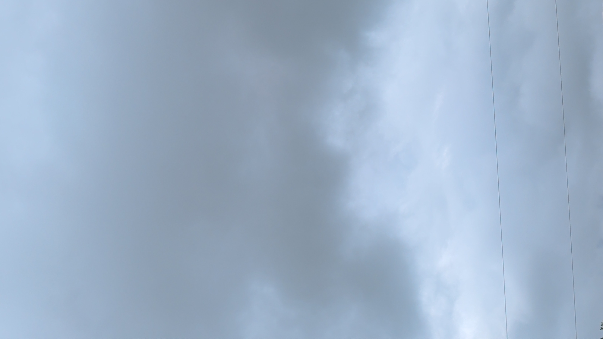 This is a vertical, eye-level, medium shot of a cloudy sky over Dorchester, Canada. The sky is predominantly light blue with some darker gray clouds towards the middle and bottom of the frame. There are two thin, horizontal lines, presumably power lines, stretched across the lower third of the image. The top two-thirds of the image is filled with a soft, hazy gradient of light blue sky. The bottom portion of the image is darker with more defined cloud formations. There are no people or discernible structures other than the power lines visible. The image appears to have been taken on an overcast day.