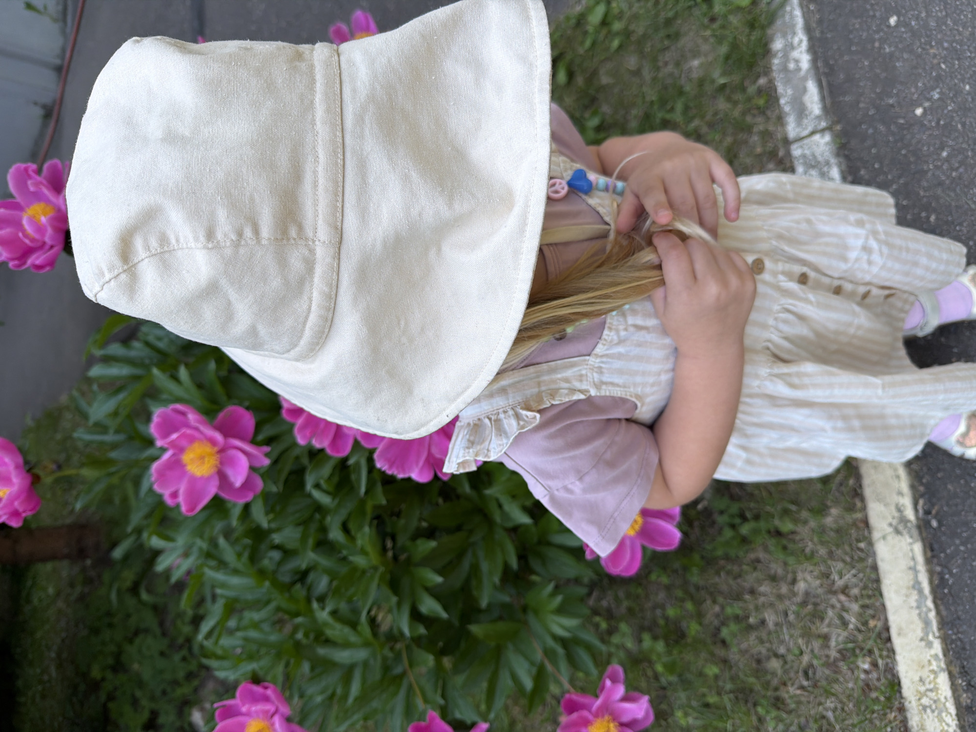 A young child wearing a sun hat, a pink t-shirt, and a striped dress is standing outdoors next to a bush with pink peonies. The child is holding their hair. The ground is covered with grass and asphalt, with a concrete border between them. The image does not provide any information about the location in Moscow, Russia.