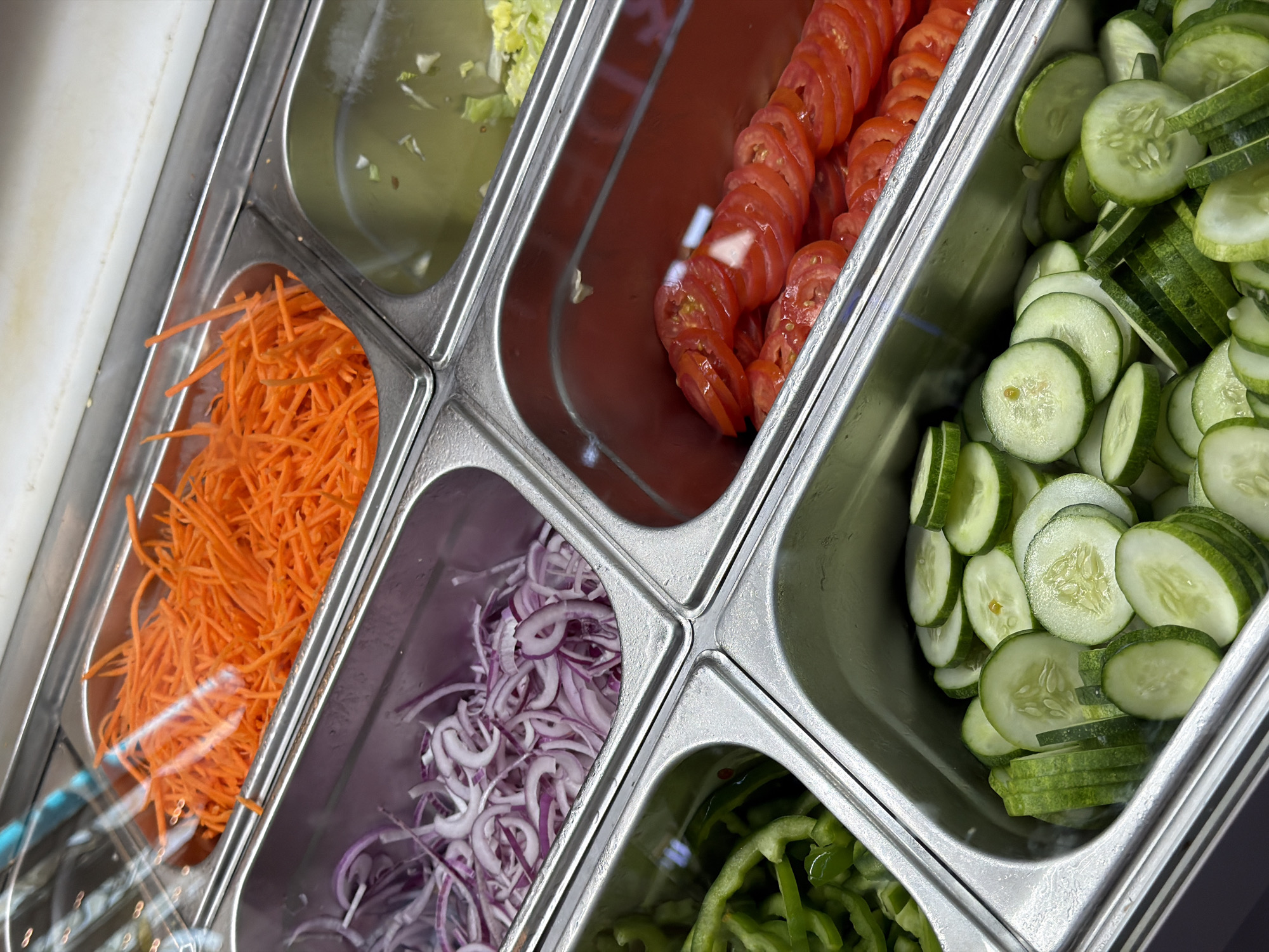 The scene depicts a close-up, overhead view of a food preparation or serving station, likely a salad bar, in ตำบล ไม้ขาว, Thailand. Six stainless steel food pans, or Gastronorm containers, are arranged within a larger metal frame.
In the top left container, a quantity of finely shredded orange carrots is present. Partially covering this container is a clear, reflective lid. Adjacent to this, in the top right, another stainless steel container holds a translucent, pale yellow liquid containing small green fragments, possibly a dressing or brine.
Below the carrots, to the left, a pan contains thinly sliced red onions. Below the red onions, to the left, another pan holds chopped green bell peppers.
Below the liquid container, to the right, a pan is filled with neatly arranged, thinly sliced red tomatoes. The interior of this pan exhibits a reddish hue. Below the tomatoes, at the bottom right of the frame, a pan contains a collection of freshly cut green cucumbers, comprising both round slices and longer, quartered pieces.
The metal surfaces of the containers and framework appear clean, with minor reflections from overhead lighting visible on the clear lids and metal. No individuals are visible in the frame, and no specific actions or interactions are occurring. The scene presents a static display of prepared salad ingredients.