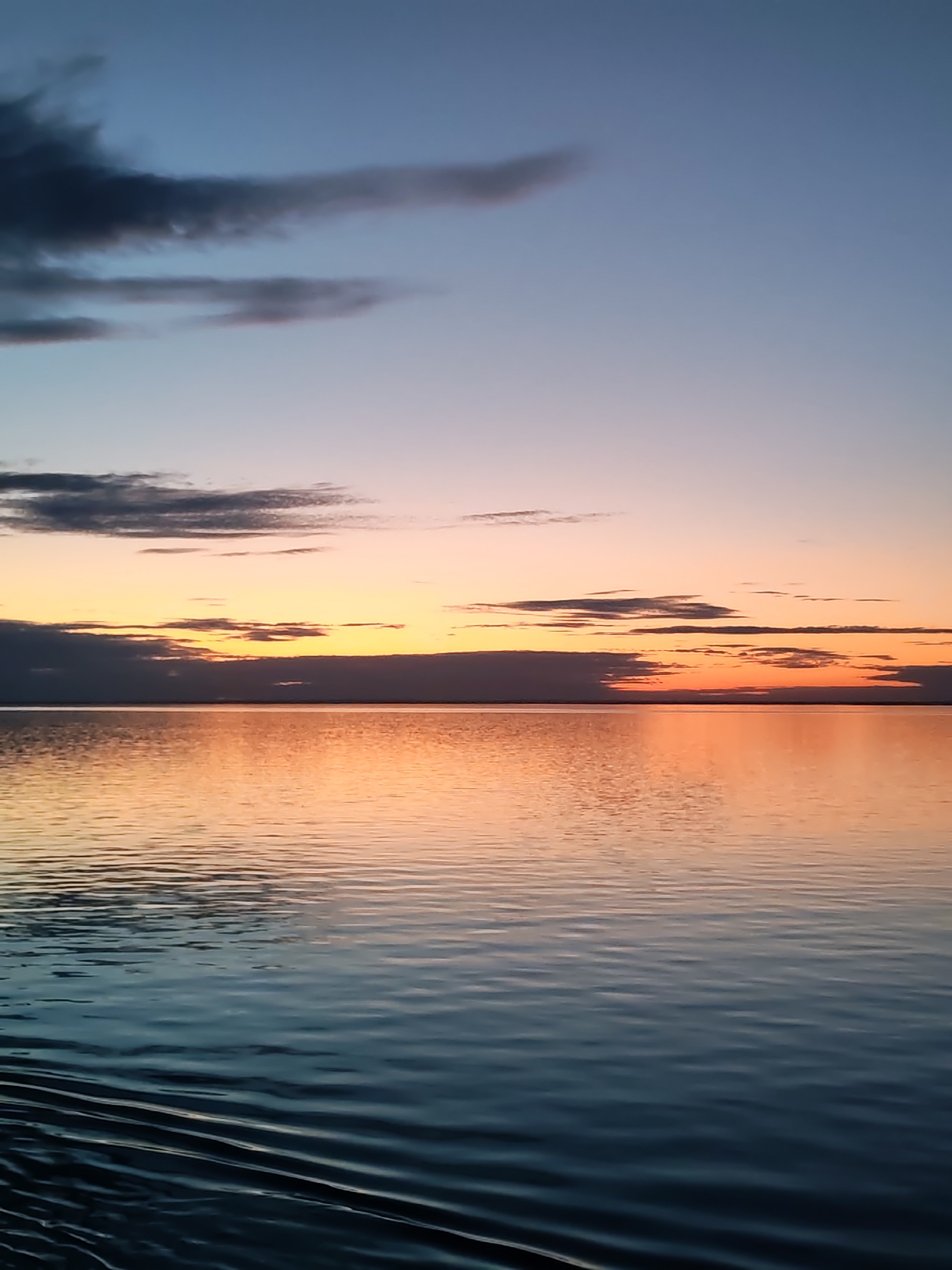 This media file presents a serene waterscape at either sunrise or sunset, characterized by a vibrant and evolving sky reflected on a vast body of water.
The scene depicts an expansive view where the sky meets the water at a clear horizon line. The upper sky transitions from a cool, pale blue-grey to warmer shades of orange, pink, and yellow closer to the horizon, indicative of the sun's position just below or within the cloud layer. Several dark, elongated clouds are scattered across the sky, with a notable band of darker clouds stretching along the horizon, partially obscuring the brightest glow of the setting or rising sun.
The water surface is calm with gentle ripples, acting as a mirror to the sky's colors. The reflection closest to the horizon glows with intense oranges and yellows, gradually blending into cooler blues, teals, and grays in the foreground, where the water appears deeper and more textured with wave patterns.
There are no people, objects, or visible text in the image. The setting suggests a large, open body of water, such as a sea or a major lake, possibly located in Russia given the context provided. The time of day is clearly sunset or sunrise, conveying a sense of tranquility and natural beauty. The weather appears partly cloudy but peaceful.