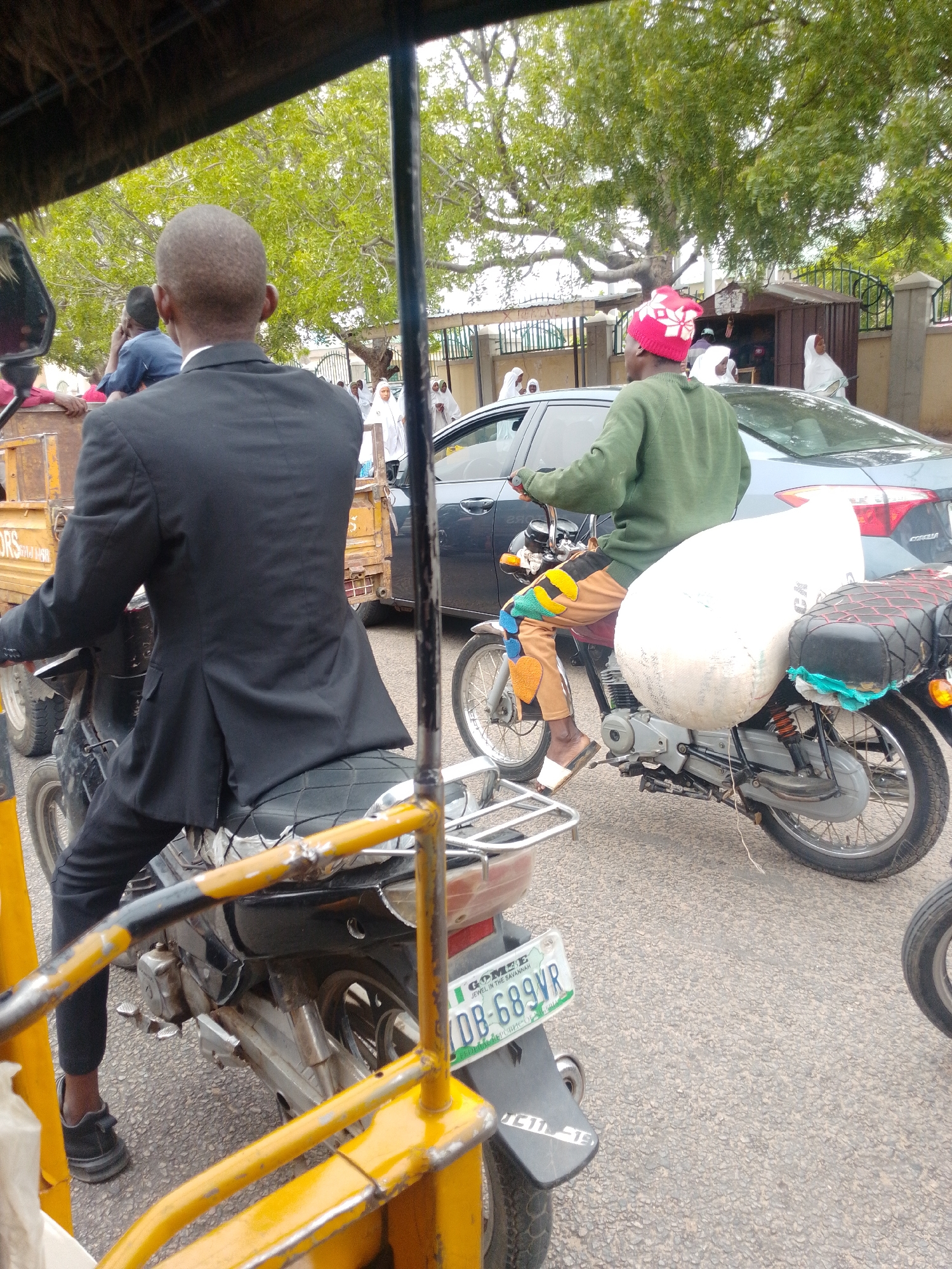 In Arawa, Nigeria, a man in a black suit rides a motorcycle, his back to the camera, navigating a street with other vehicles and pedestrians. To his right, another man on a motorcycle, wearing a green sweater and a red and white patterned hat, carries a large white sack. Behind them, a grey sedan and a yellow truck are visible. Pedestrians in white attire can be seen in the background, near a building with a dark metal fence. The environment appears to be an urban setting with trees and paved roads. The license plate on the rear of the first motorcycle is partially visible and reads "DB-689VR".