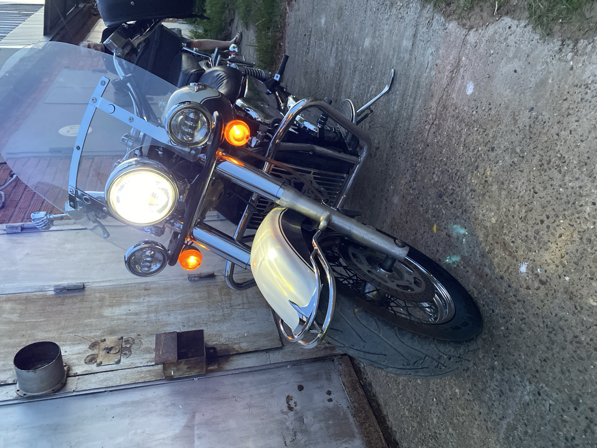 The image shows the front of a parked motorcycle, with its headlight on. The motorcycle has a windscreen, chrome details, and lit turn signals. It is parked next to a weathered garage door. The surface underneath the motorcycle is paved. It is unclear if the location is in Chaikovsky, Russia.