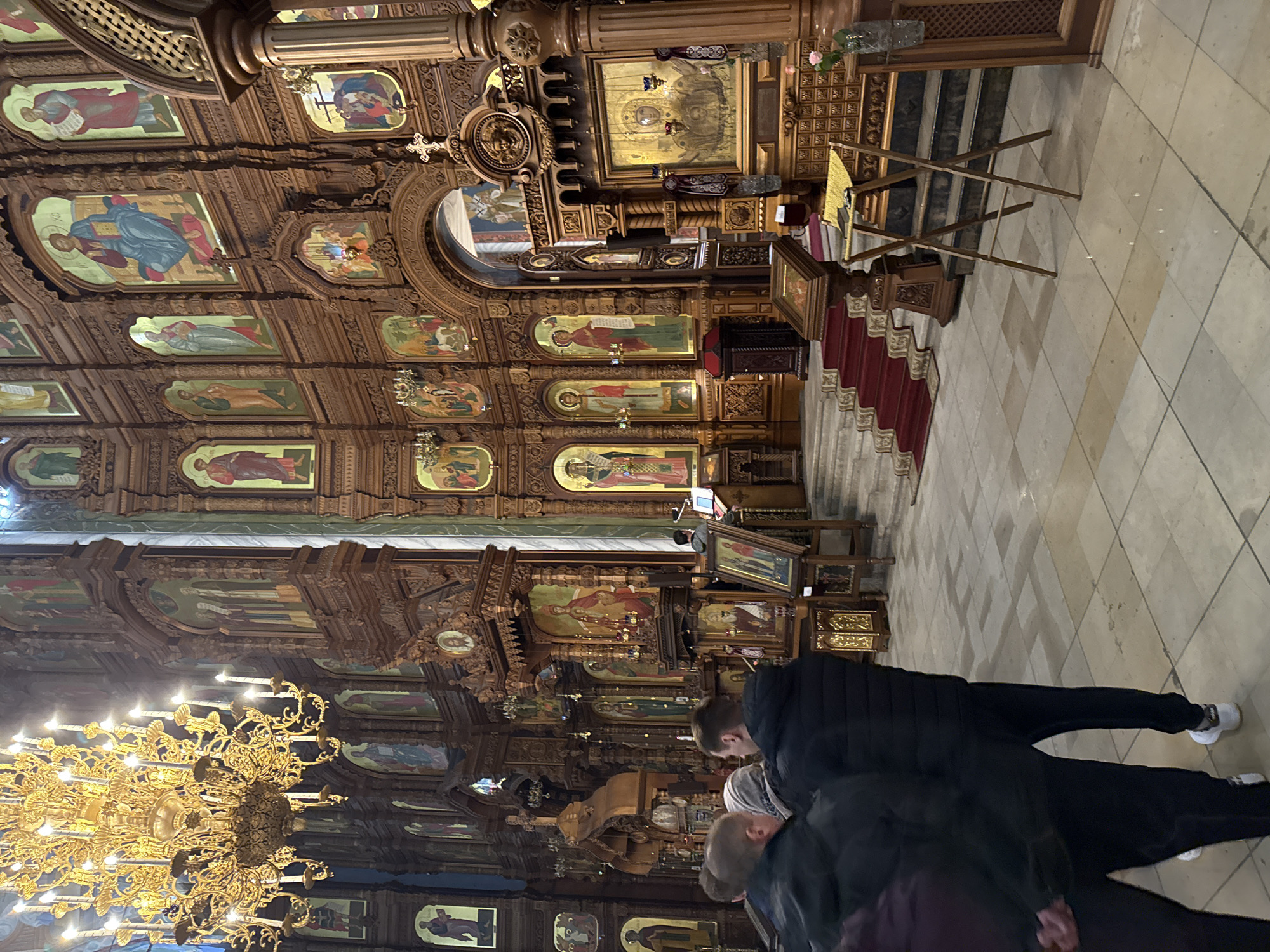 The image shows an interior view of a Russian Orthodox church. The walls are adorned with numerous icons and ornate wooden carvings. A large, elaborate chandelier hangs from the ceiling. Several people are present, standing near the wall with the icons, seemingly observing the artwork. The floor is tiled. The location is likely in Nizhny Novgorod, Russia, given the architectural style and religious iconography.